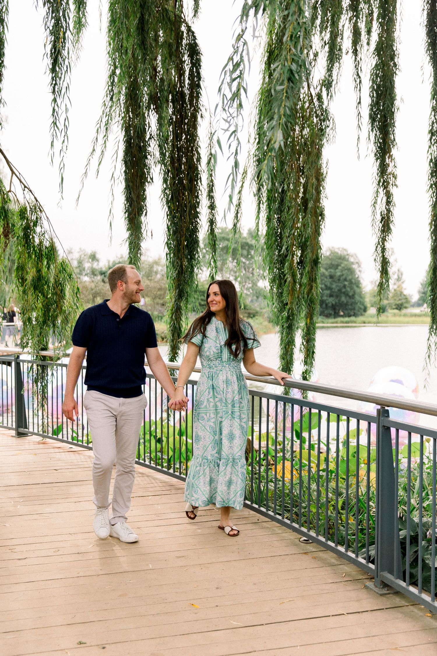 Couple holding hands and smiling on a wooden boardwalk under a weeping willow tree near a serene lake.