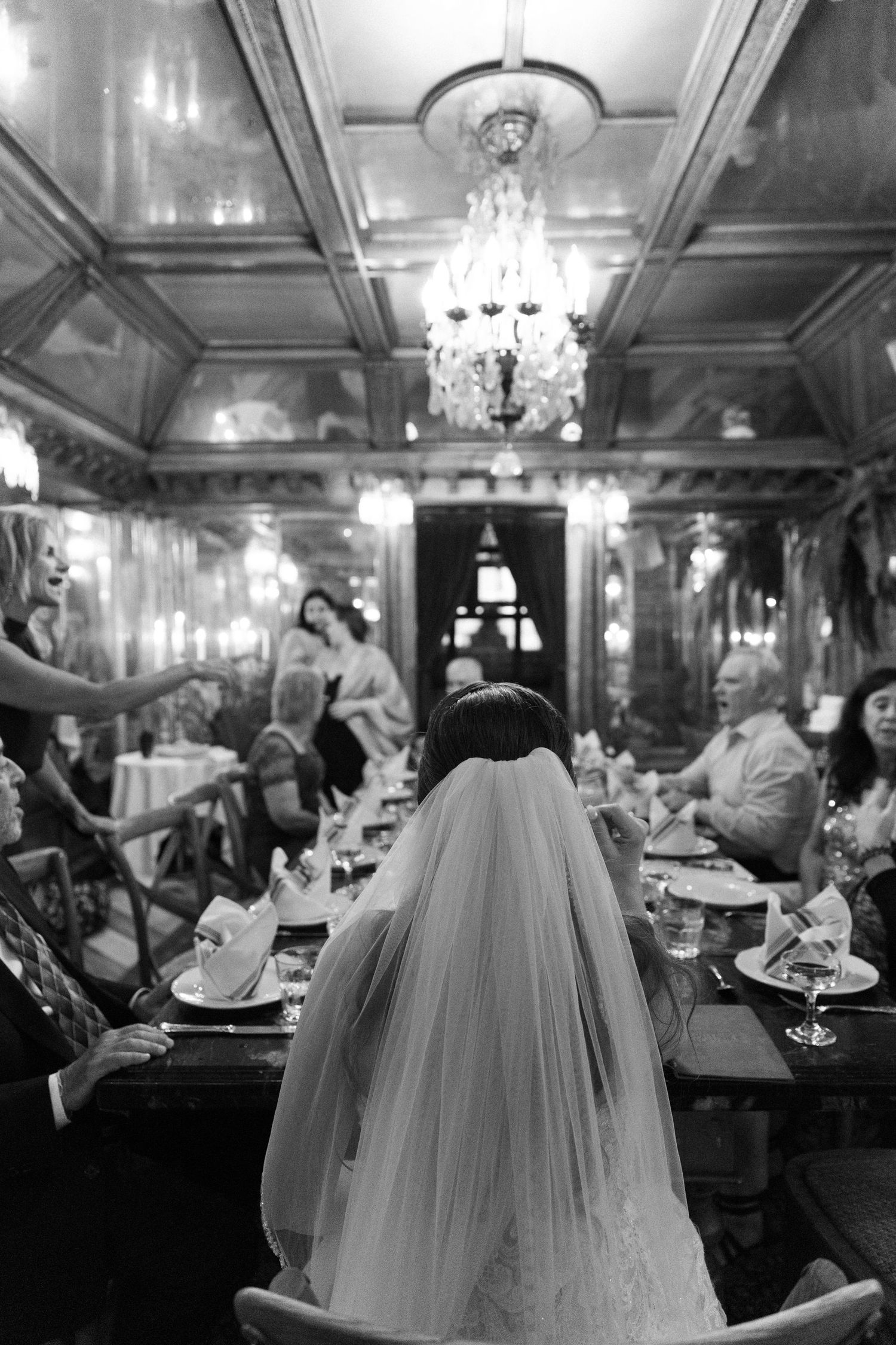 Bride in veil seated at ornate candlelit wedding reception dinner table, black and white.