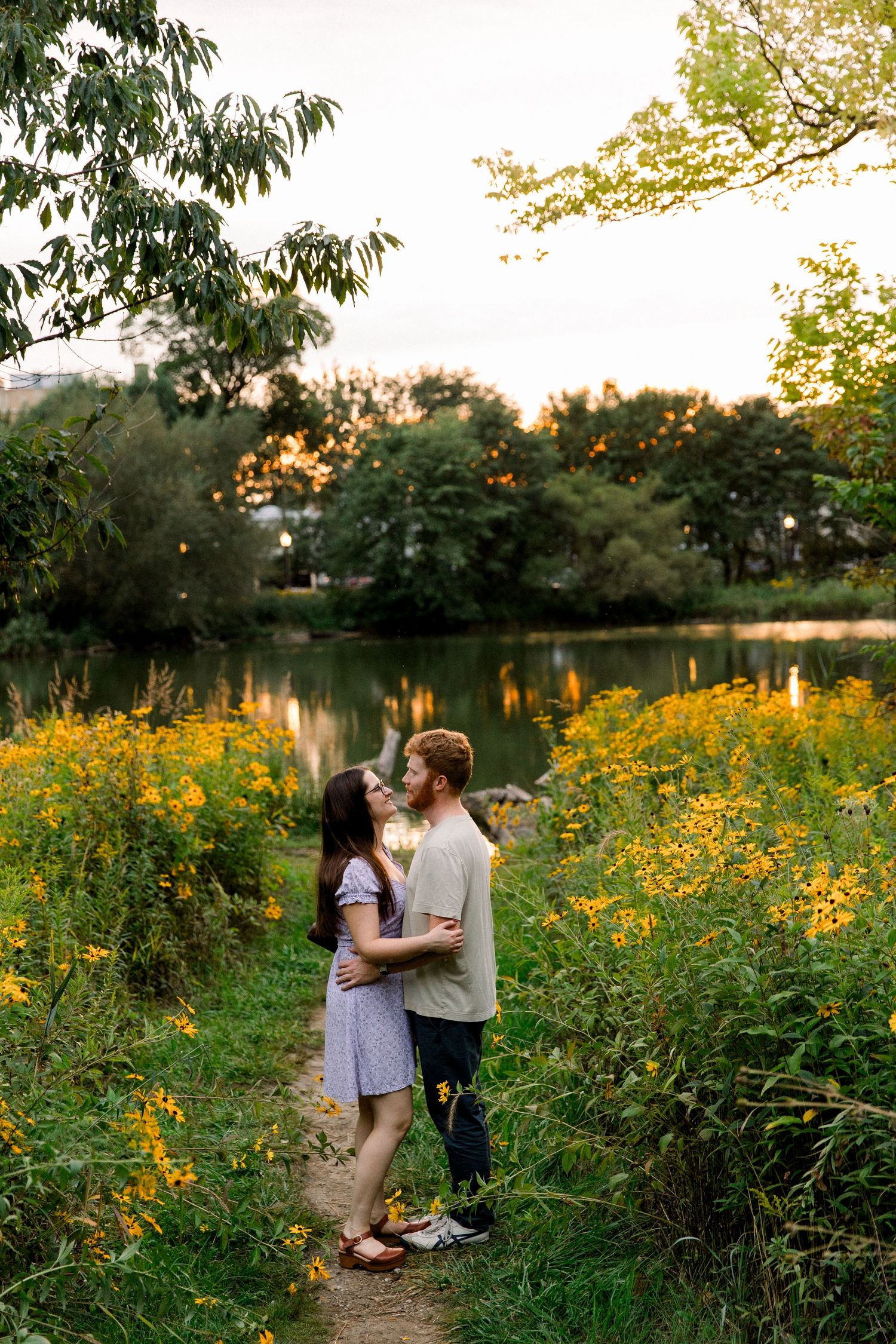 Couple embracing among yellow wildflowers by a serene pond at golden hour sunset in a lush green park.