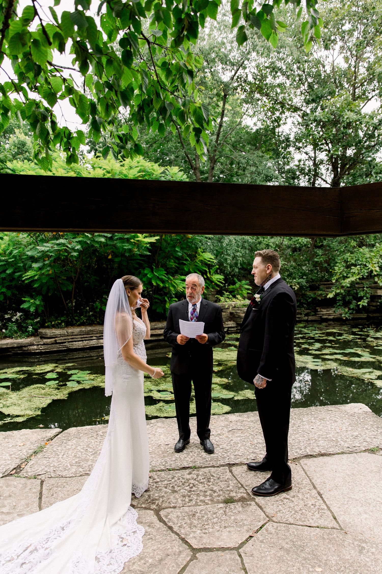 Bride and groom exchange vows outdoors by a lily pond with an officiant in a lush garden.