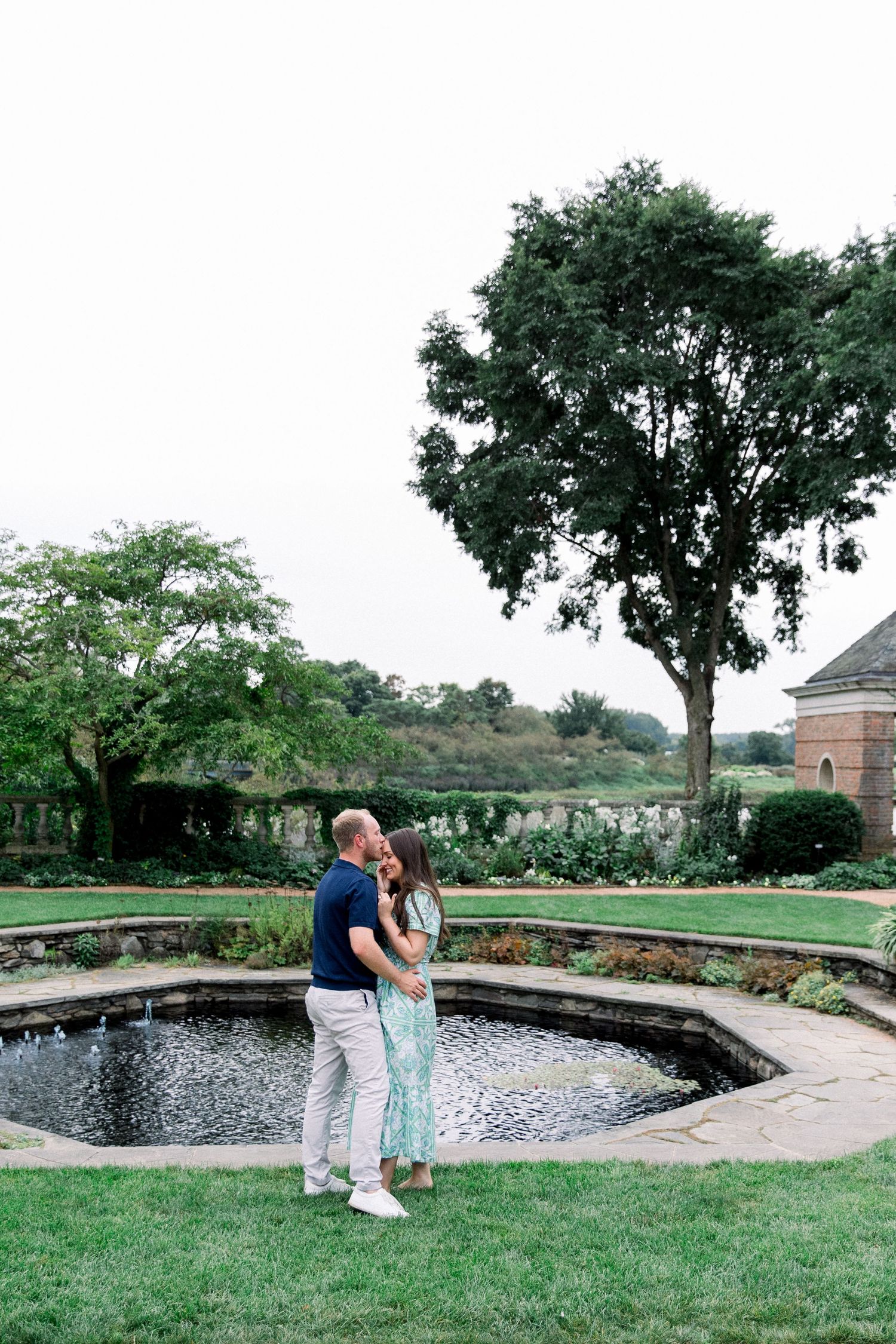 Couple embracing near a stone fountain in a lush garden with tall trees and manicured hedges.