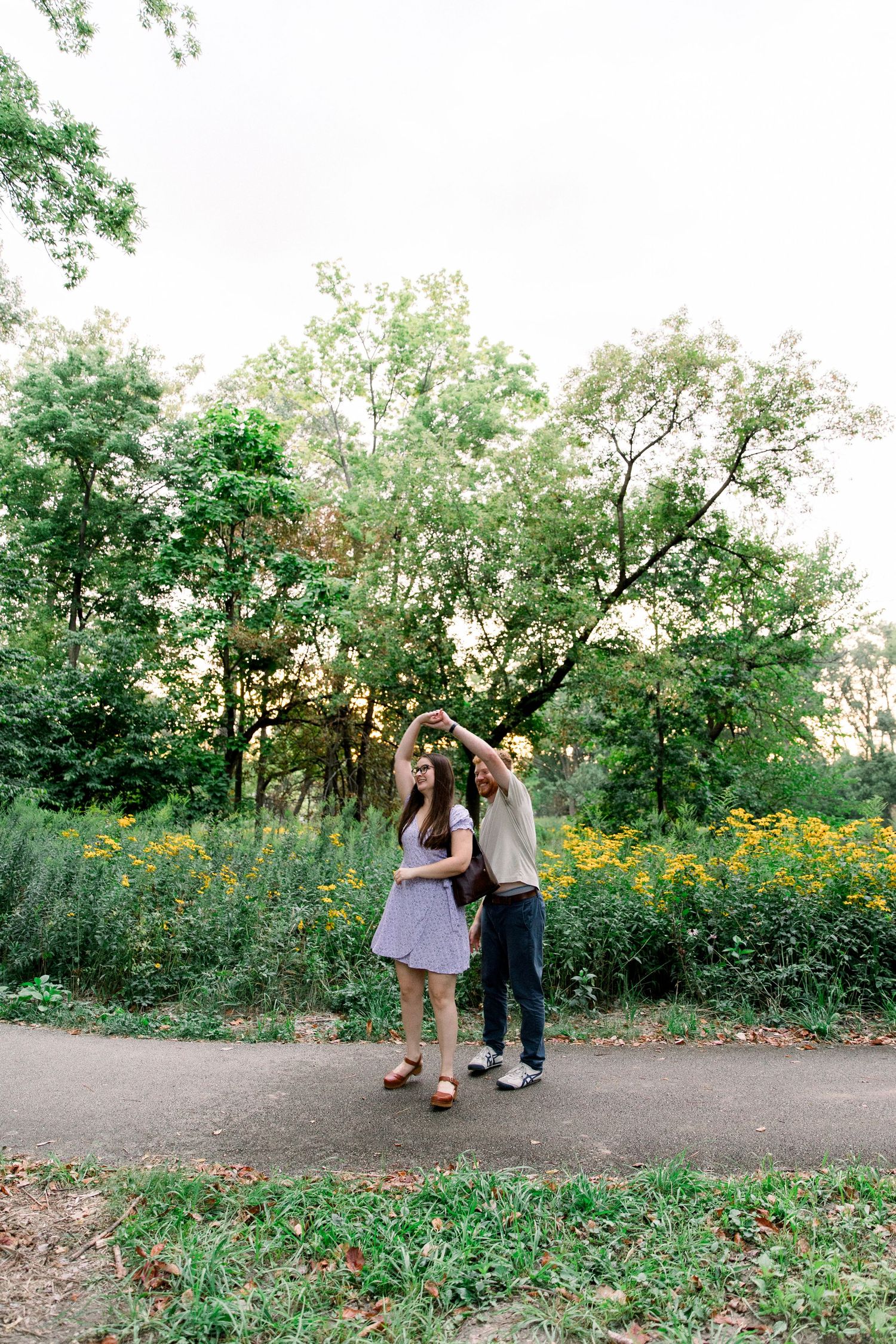 Happy couple dancing on a park path surrounded by yellow wildflowers and green trees at golden hour.