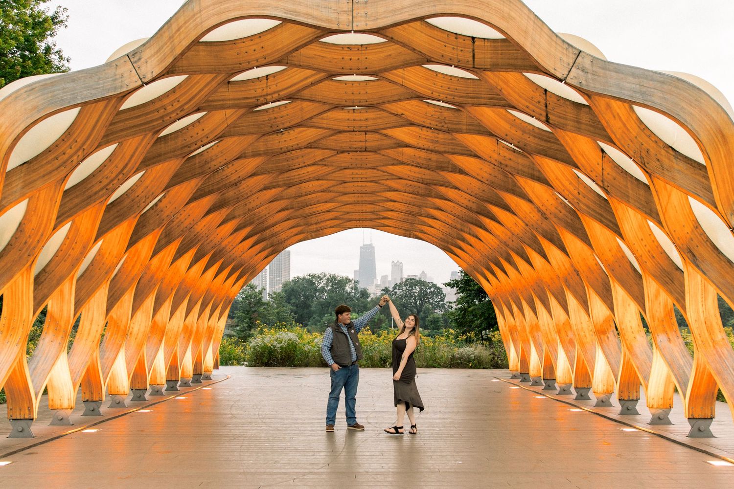 Couple standing under a stunning wooden lattice tunnel arch with warm orange tones and city skyline in background.