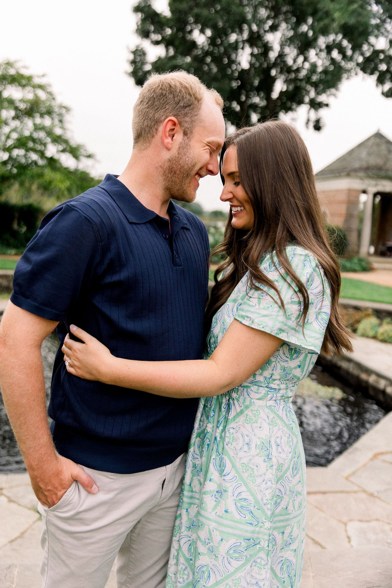 Happy couple embracing and smiling at each other outdoors, man in navy polo and woman in green floral dress near garden pond.