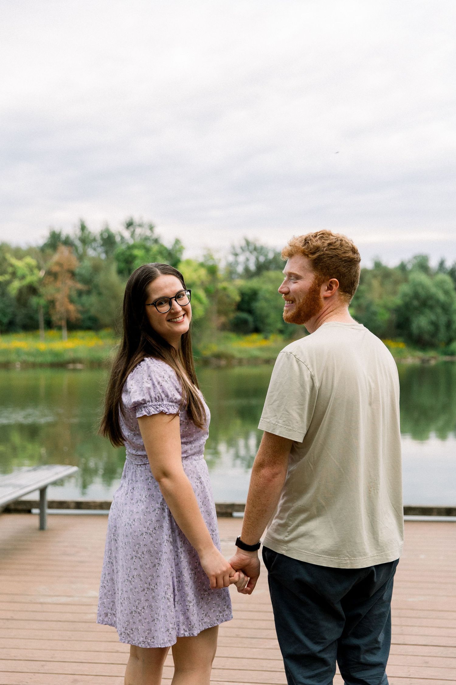 Couple holding hands on a lakeside dock, woman in floral dress smiling, lush green trees reflected in calm water.