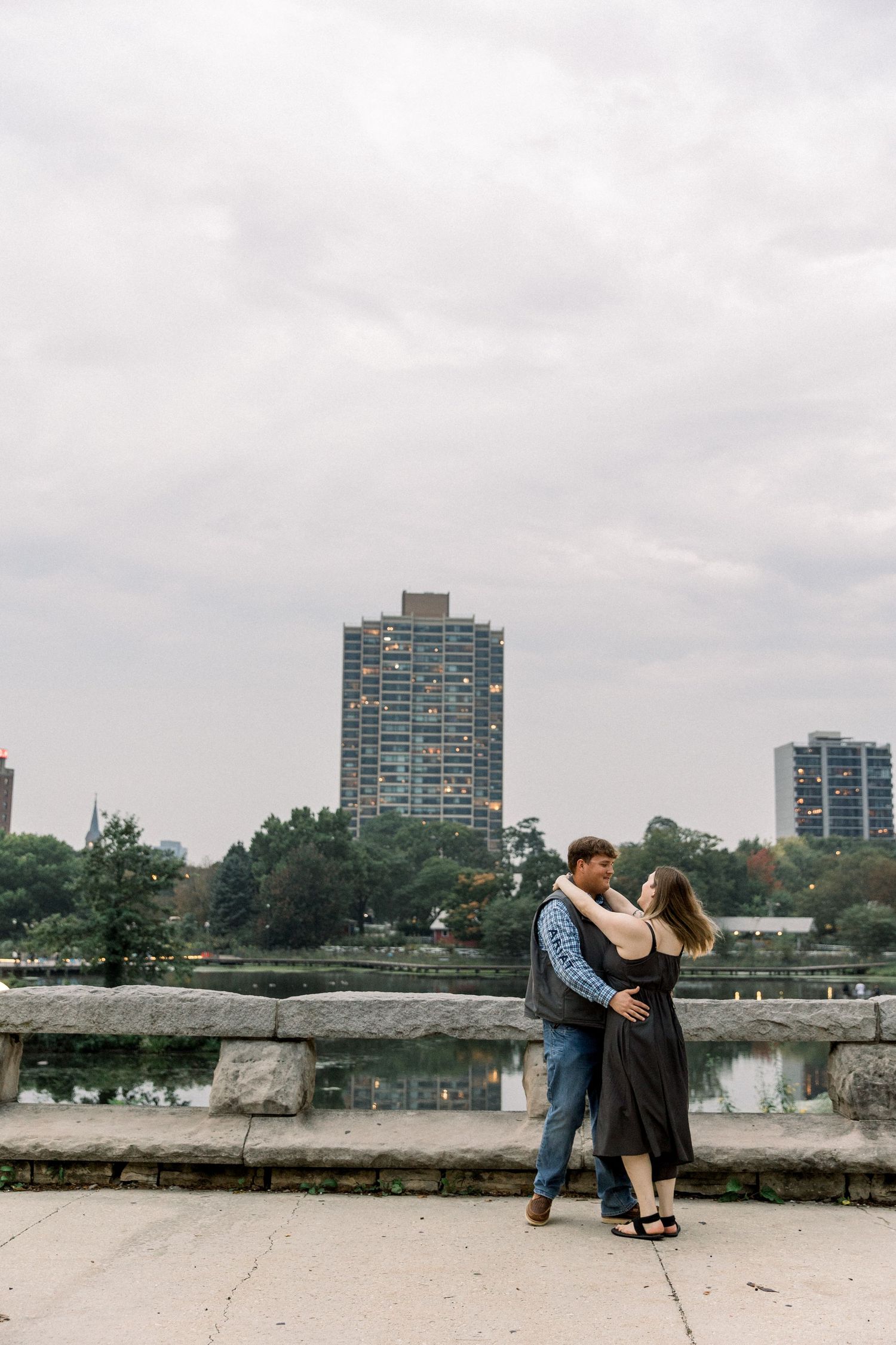 Couple embracing romantically on a stone walkway with city skyline and lake in the background on a cloudy day.