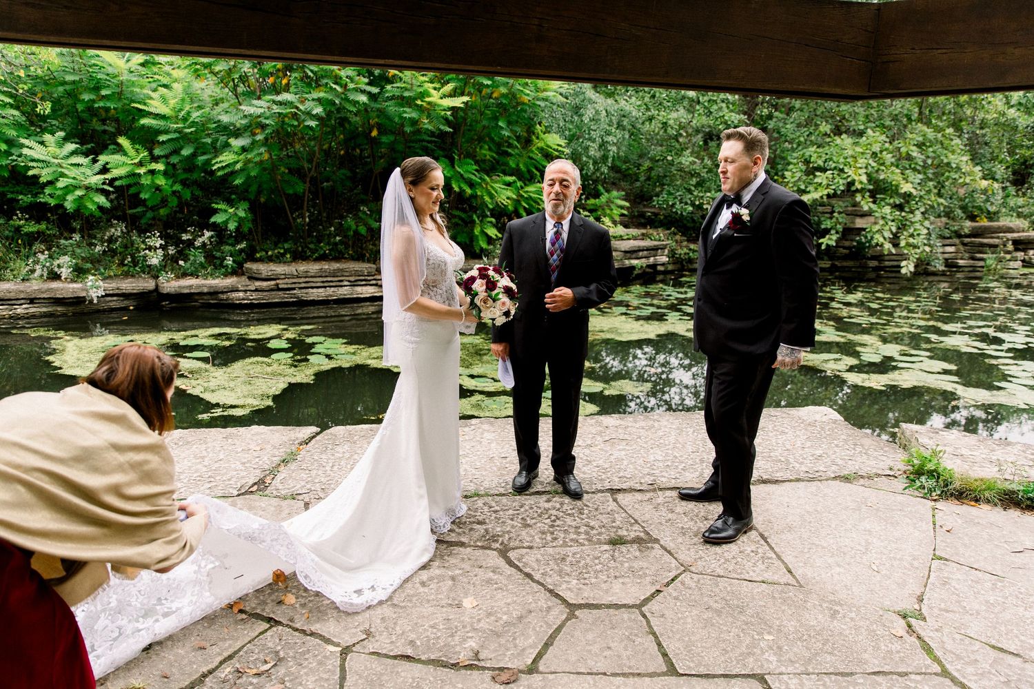 Outdoor wedding ceremony by a lily pond with bride, groom, and officiant under pavilion.