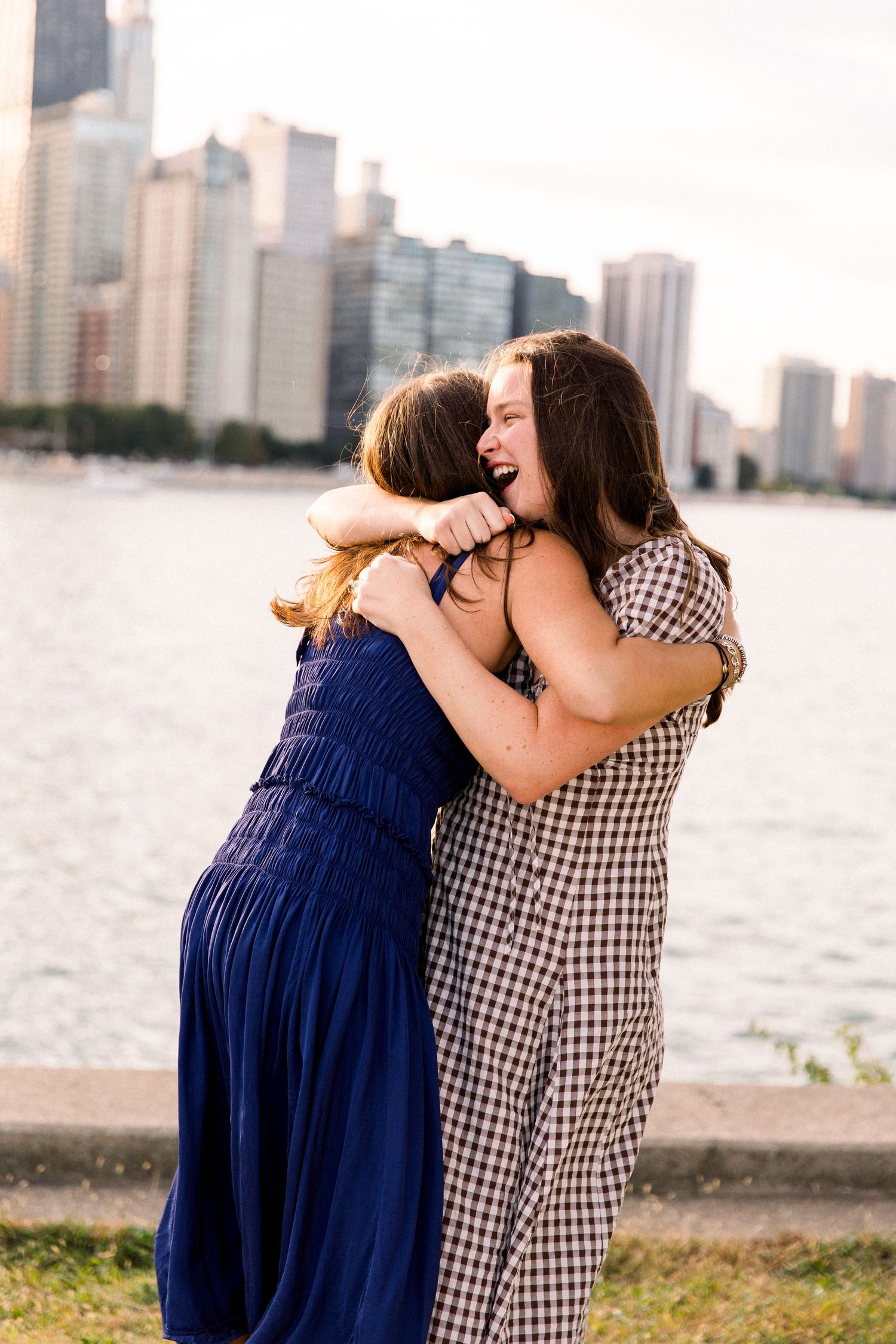 Two women hugging and laughing by a waterfront with a city skyline backdrop, one in blue dress, other in gingham.