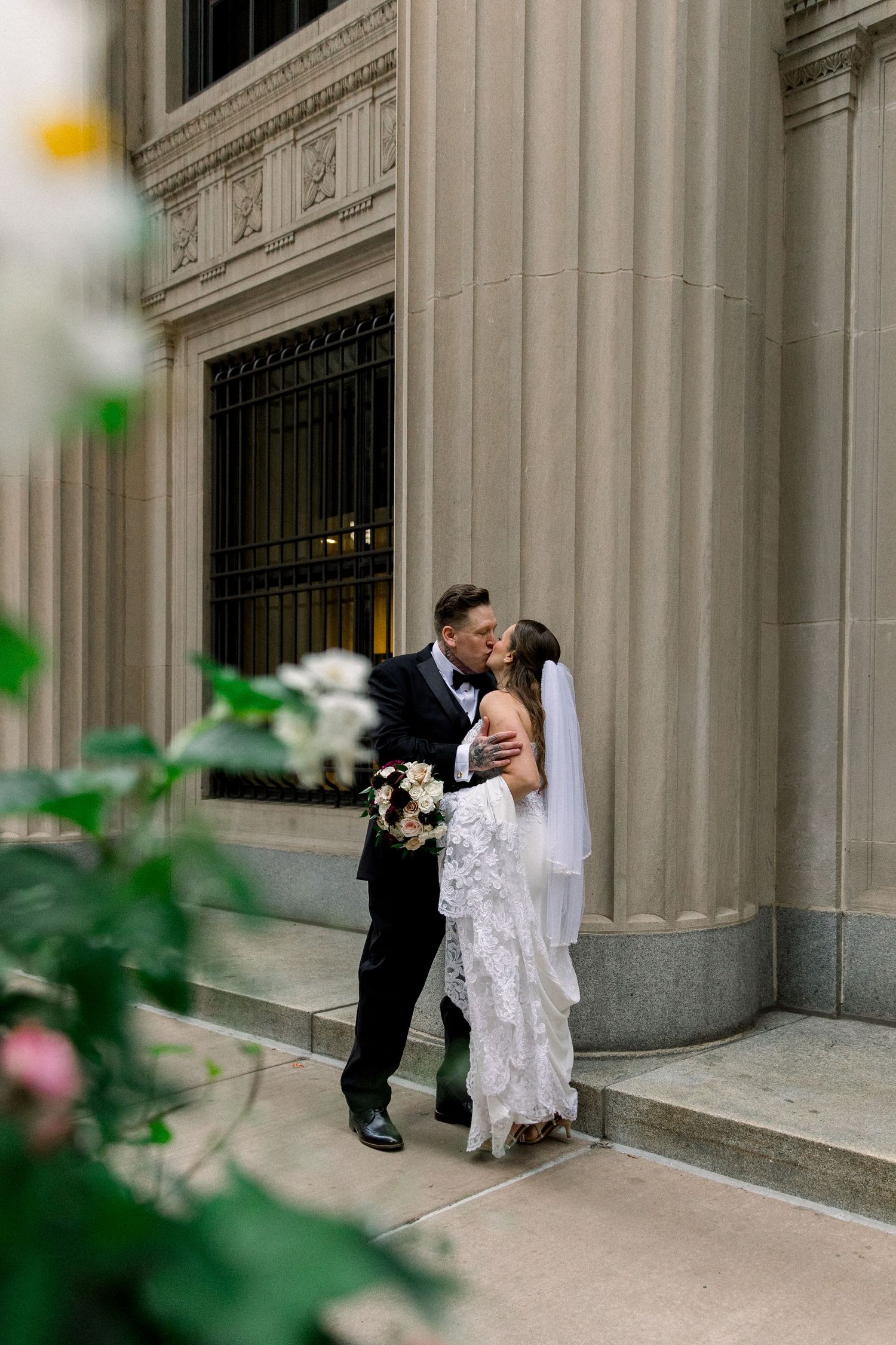 Groom kissing bride beside grand stone columns with lace wedding dress and veil.