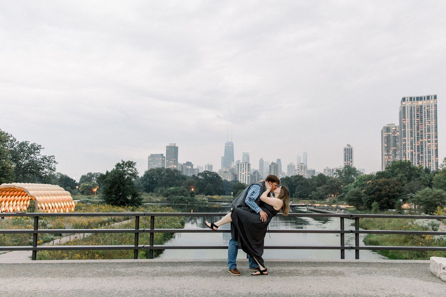 Couple sharing a romantic dip kiss on a bridge with Chicago skyline backdrop on a cloudy day.