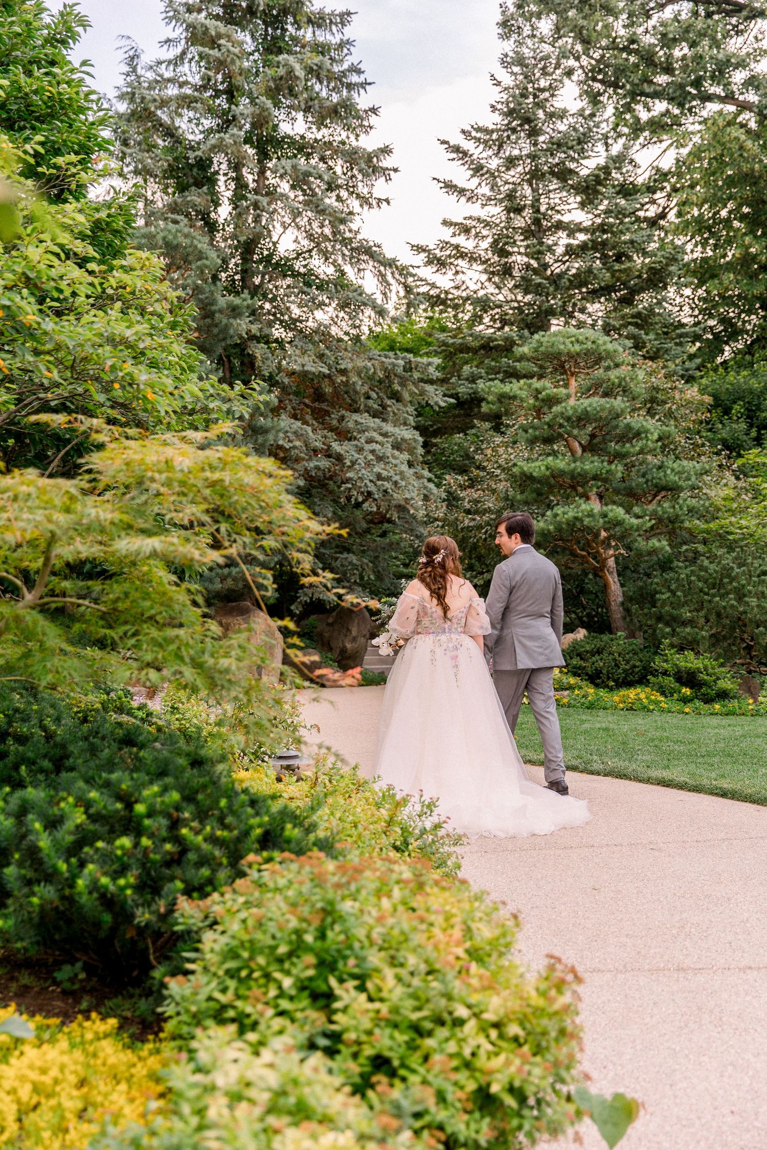 Bride and groom walk hand in hand along a garden path surrounded by lush greenery on their wedding day.