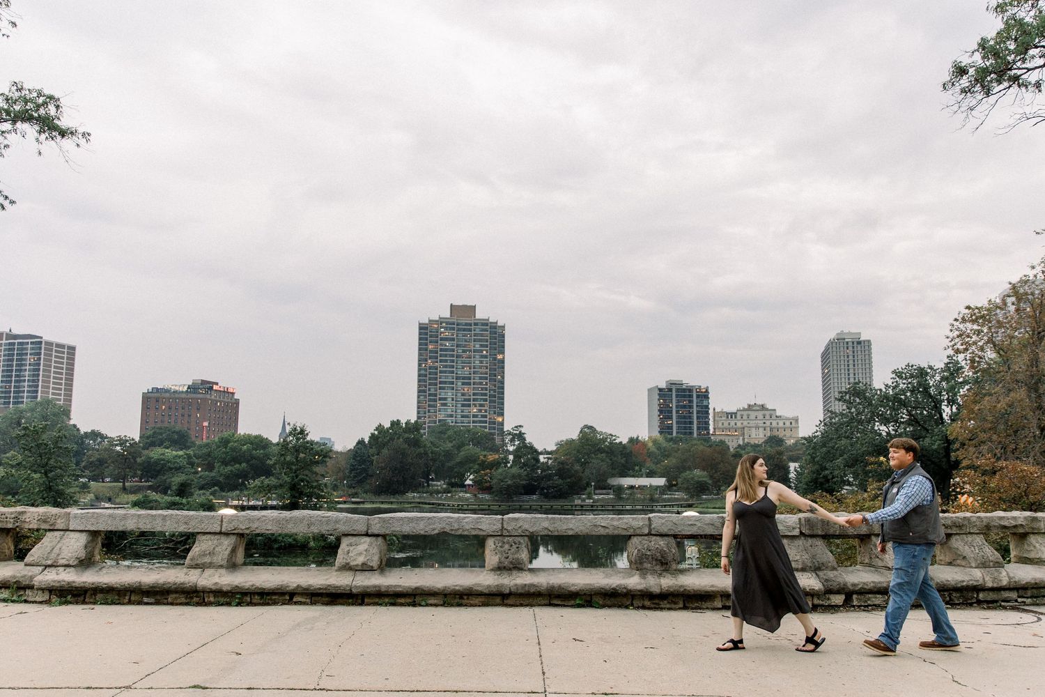 Couple holding hands walking along a stone bridge in an urban park with city skyline in the background.