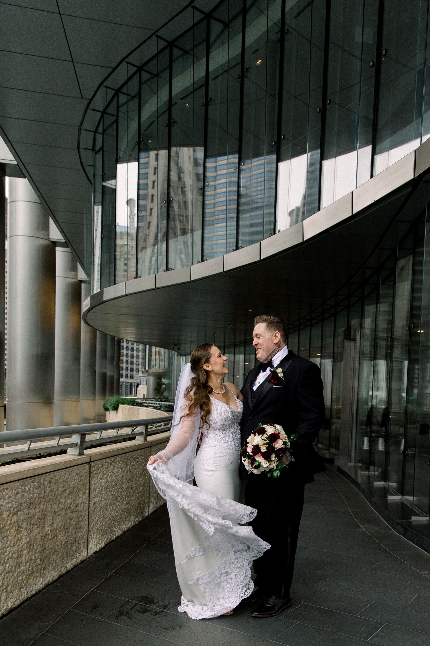 Bride and groom share a romantic moment outside a modern glass building, her lace veil flowing in the wind.