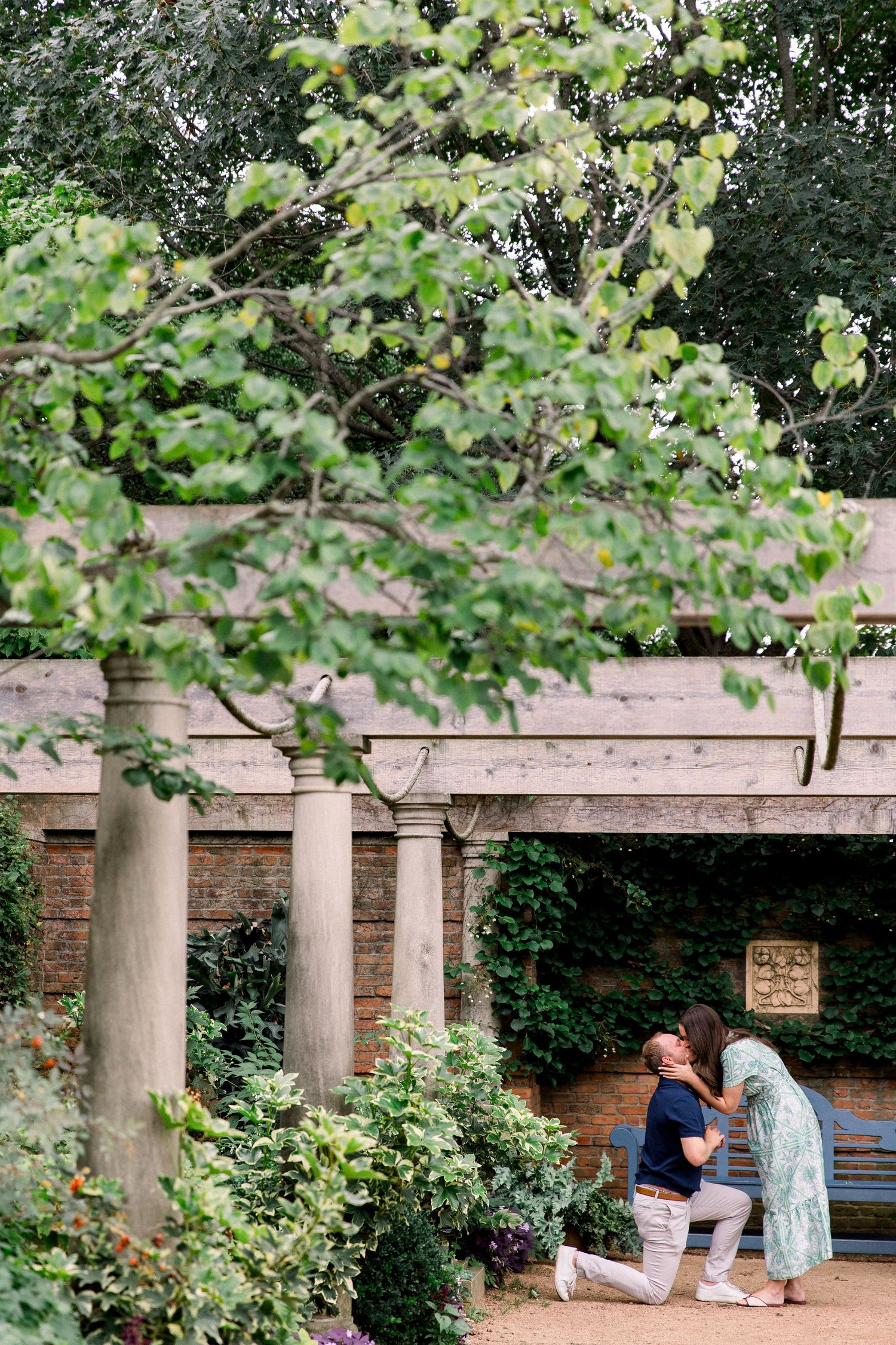 Man proposing on one knee to woman in lush garden with stone columns and ivy-covered walls.