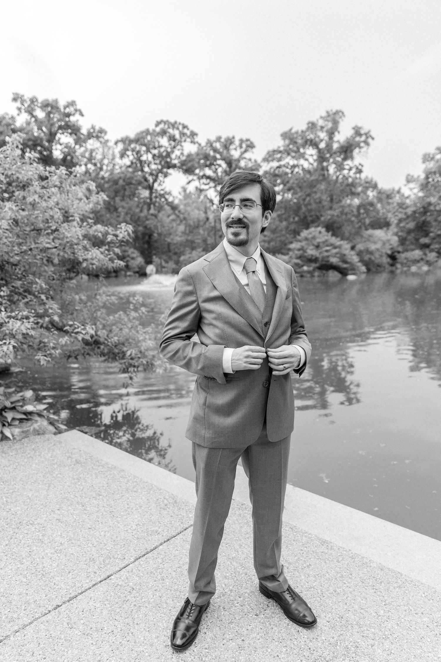 Black and white photo of a smiling groom in a suit buttoning his jacket outdoors near a serene pond.