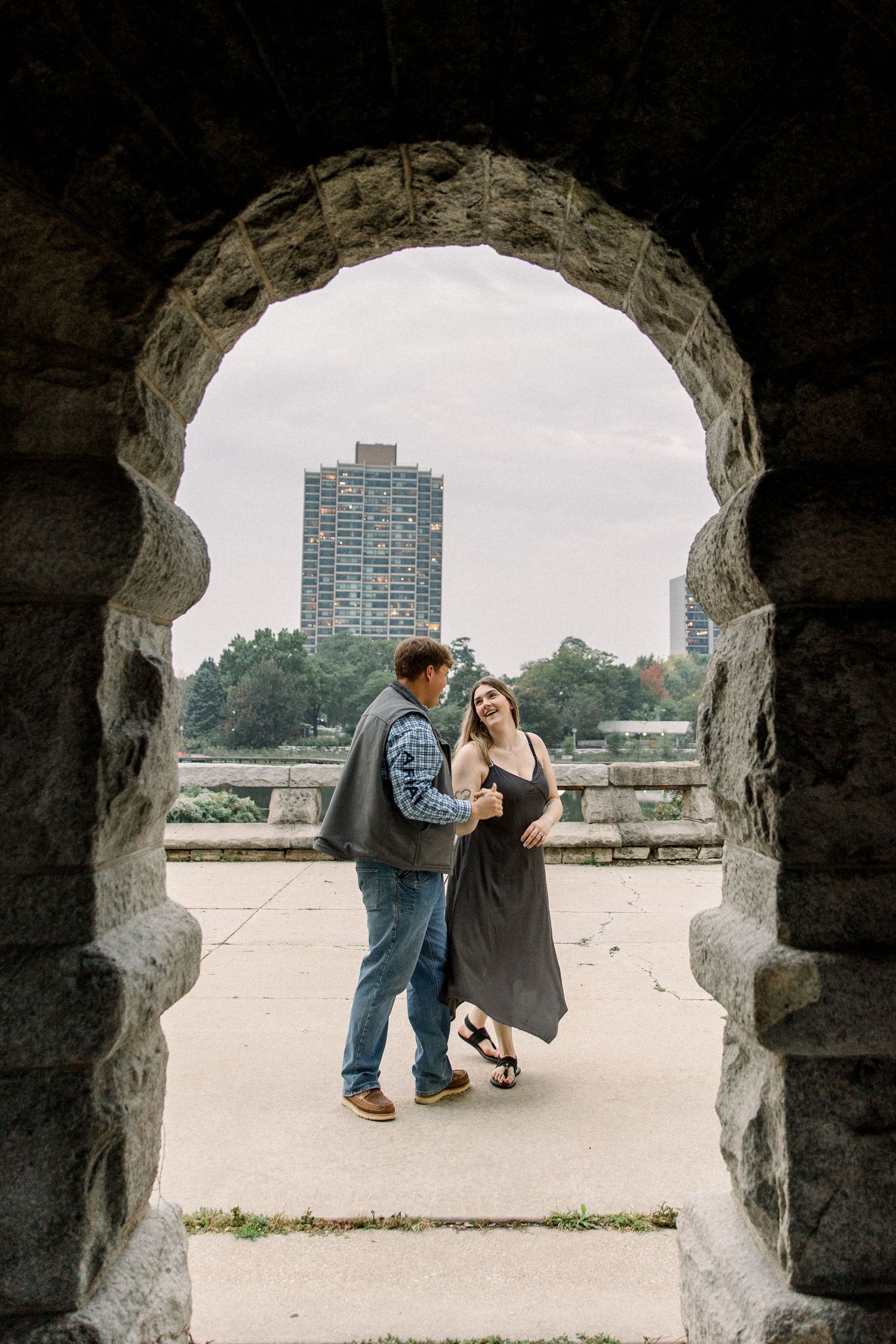 Couple dancing romantically framed by a stone arch in a park with a city skyline in the background.