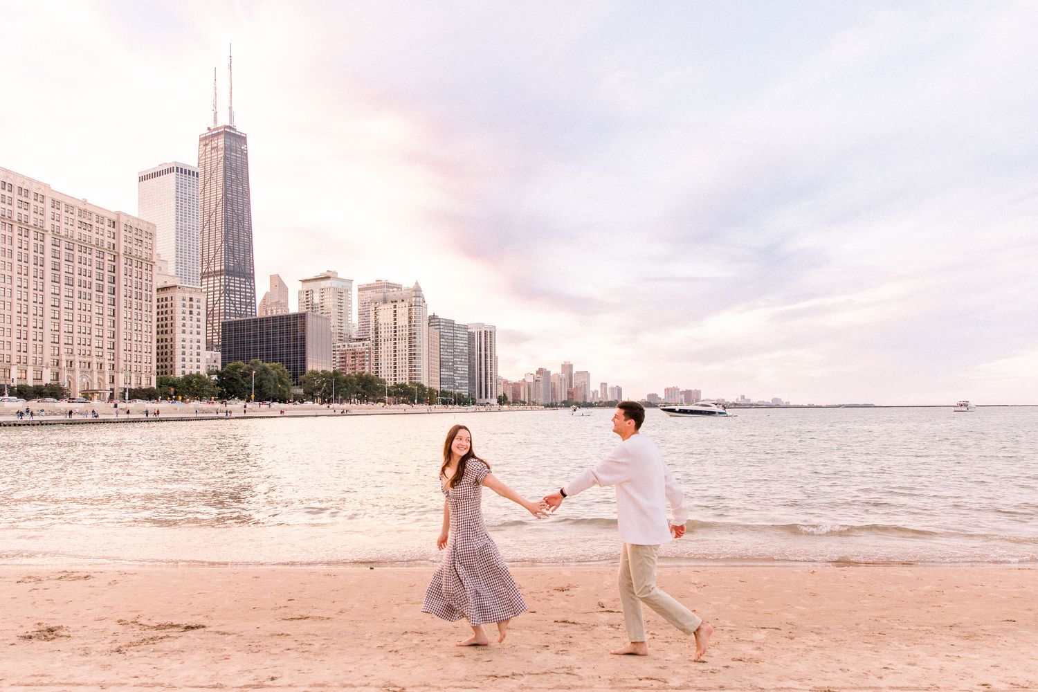 Couple holding hands walking on Chicago beach with city skyline and John Hancock Center in background at sunset.