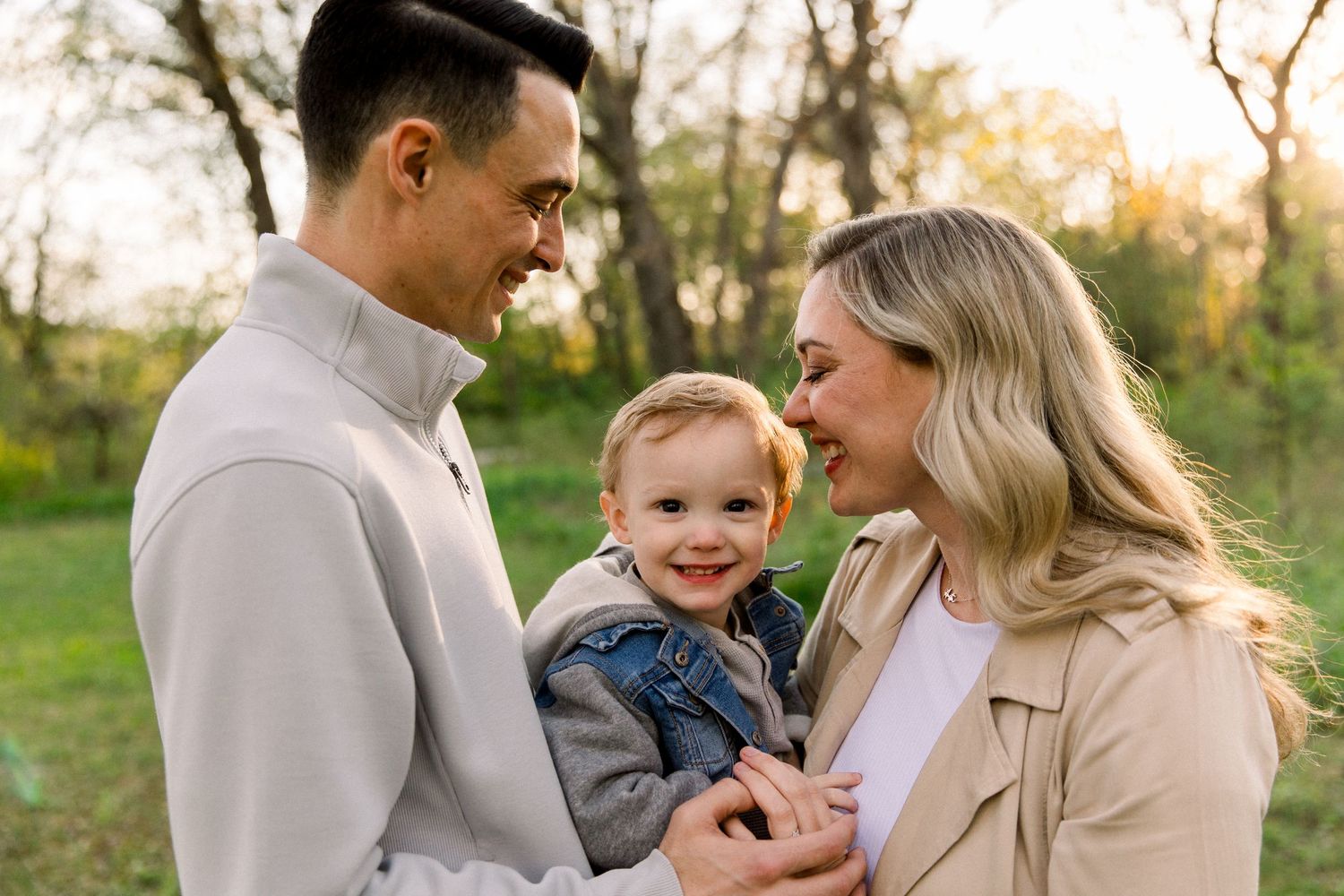 A happy young family with a smiling toddler poses outdoors in a lush green park during golden hour.