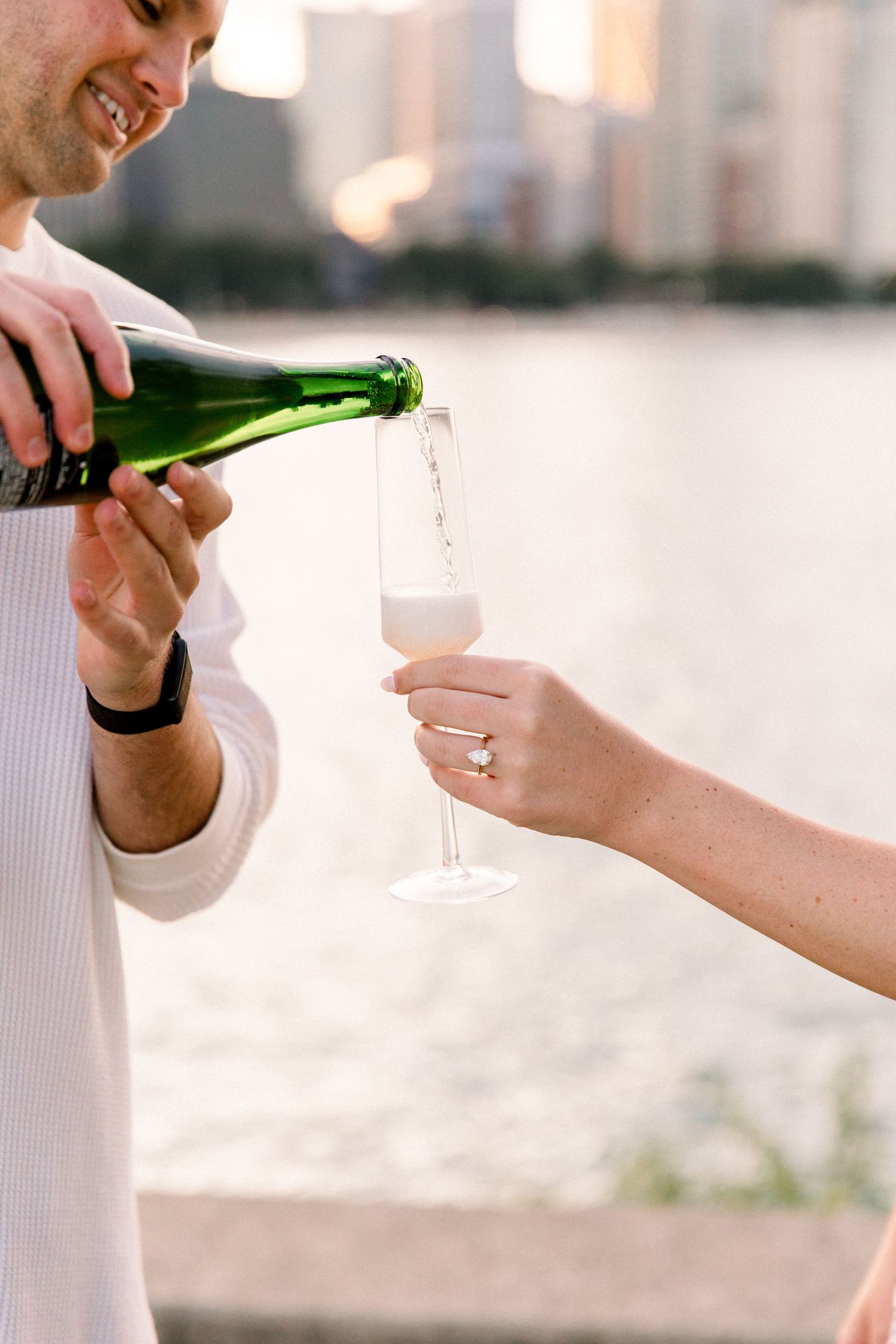 Man pouring champagne into a flute held by a woman wearing an engagement ring, celebrating by a waterfront.