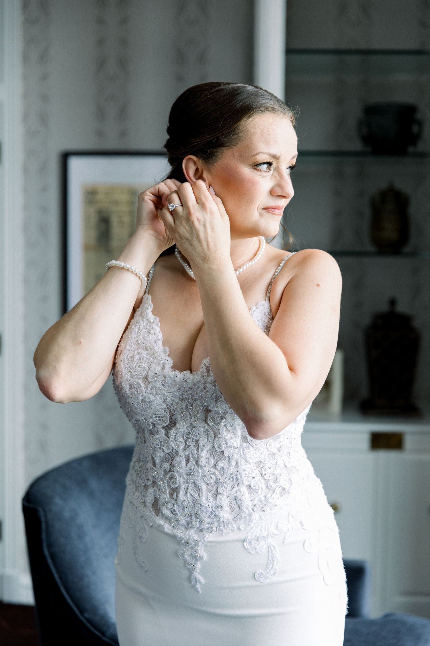 Bride in lace wedding dress adjusting earrings, wearing pearl bracelet and necklace, smiling in elegant room.