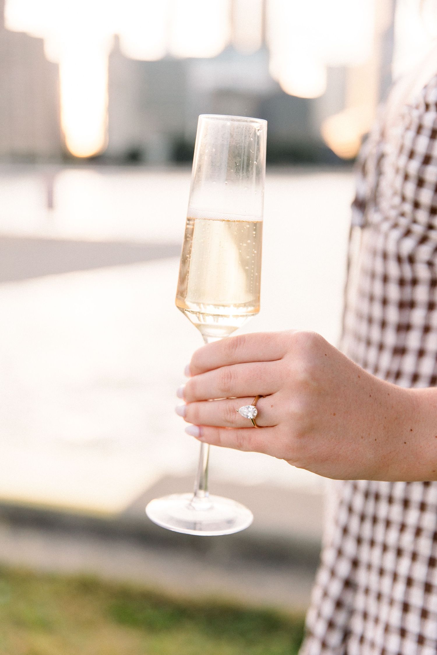 Close-up of woman's hand holding champagne flute with diamond engagement ring against blurred city backdrop.