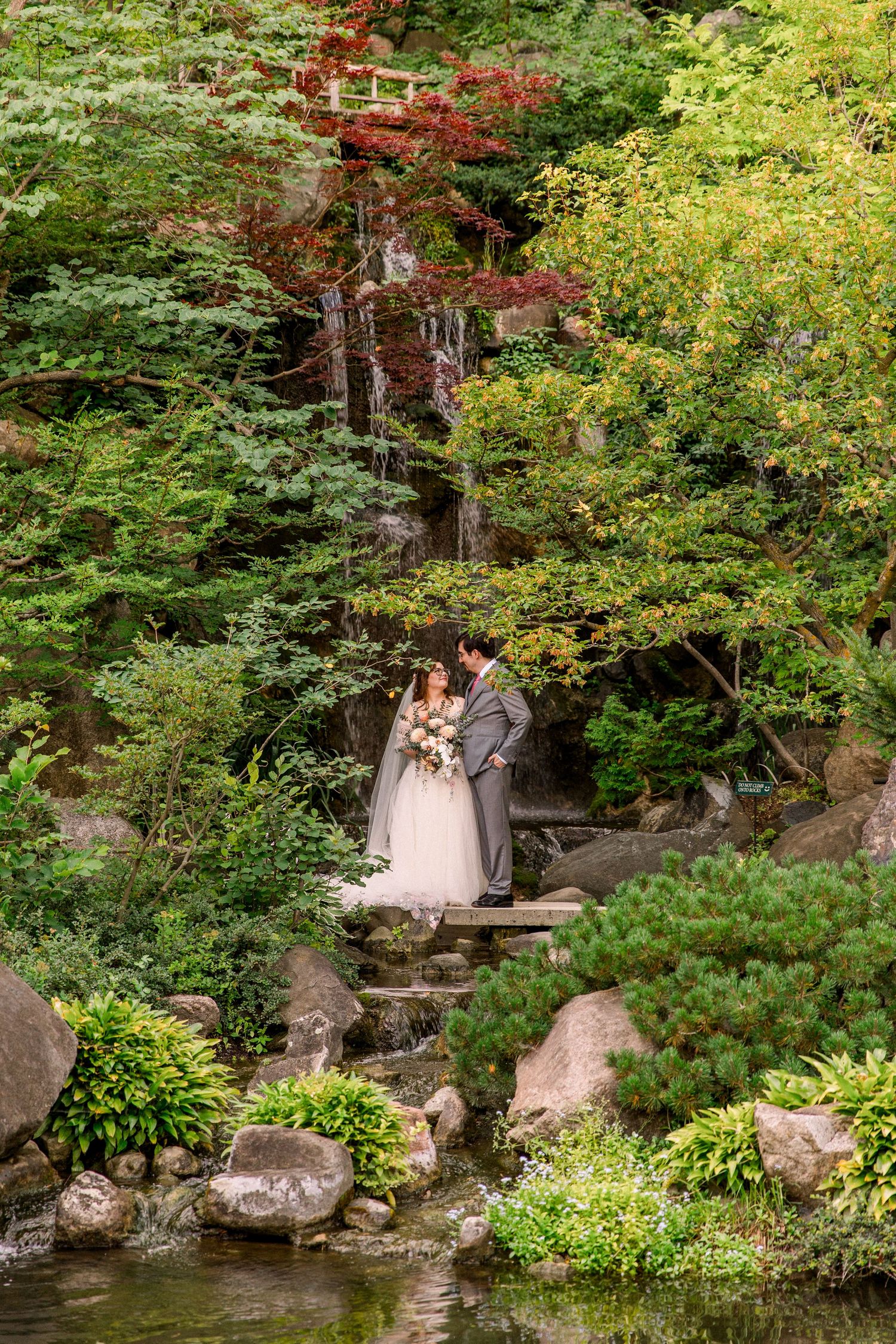 Bride and groom stand on stone steps surrounded by lush greenery and rocks in a serene Japanese garden setting.