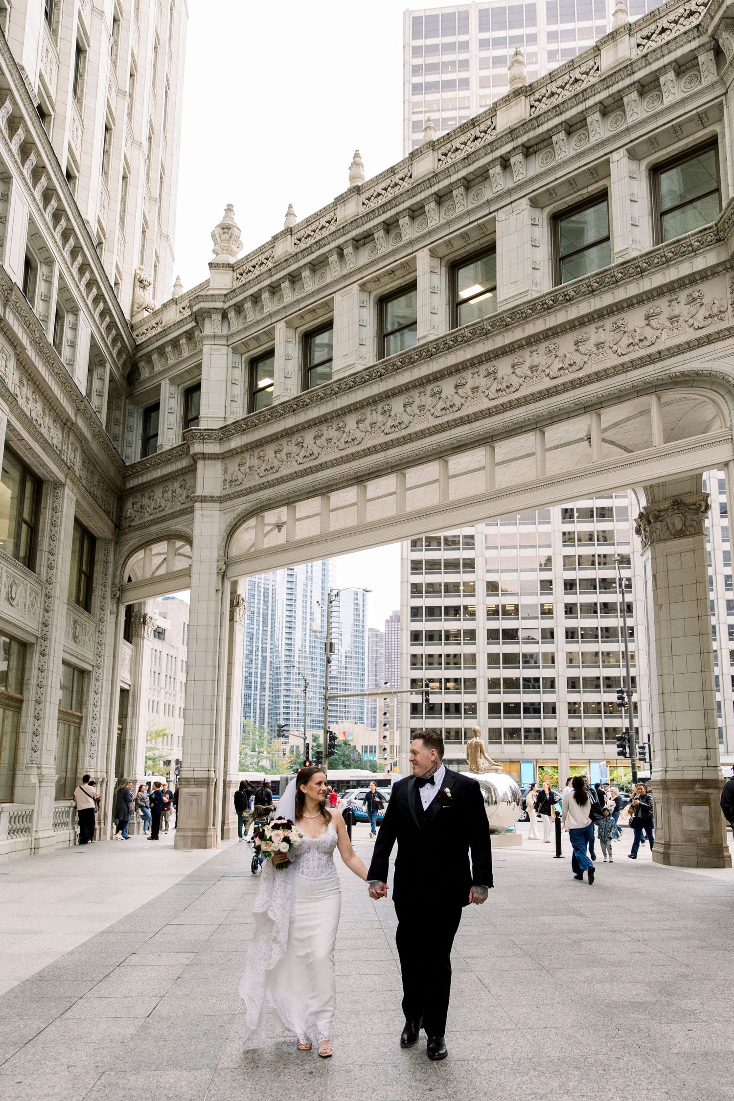 Bride and groom walk hand-in-hand beneath ornate stone arch in downtown Chicago on their wedding day.