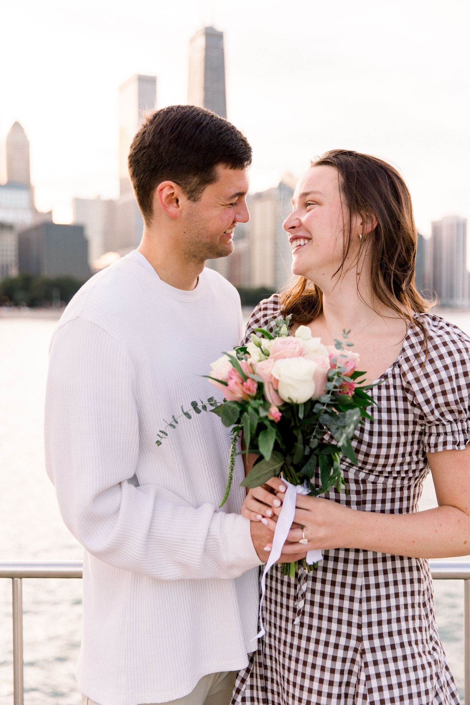A smiling couple shares a romantic moment holding a bouquet of pink and white roses with a city skyline backdrop.