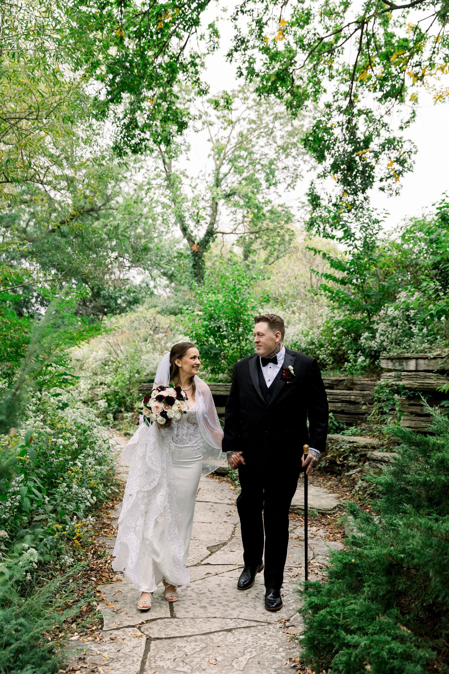 Bride and groom walk hand-in-hand along a stone garden path, surrounded by lush greenery on their wedding day.