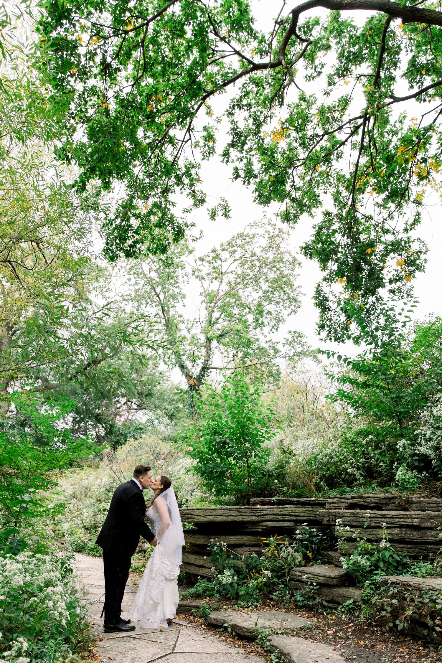 Bride and groom share a kiss on a stone path surrounded by lush green trees in an outdoor garden wedding setting.