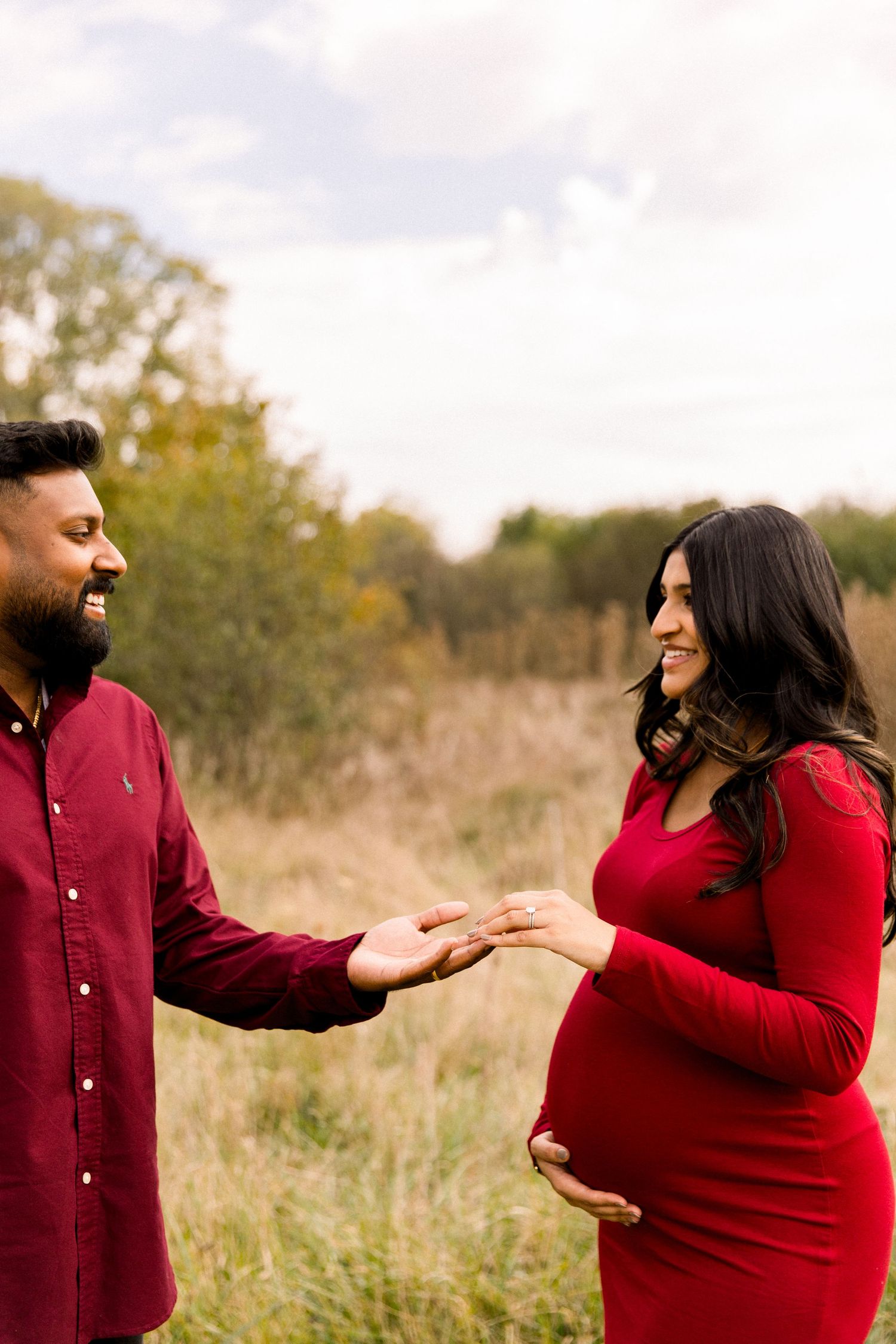 Smiling pregnant couple in red outfits holding hands in a field during a maternity photoshoot.