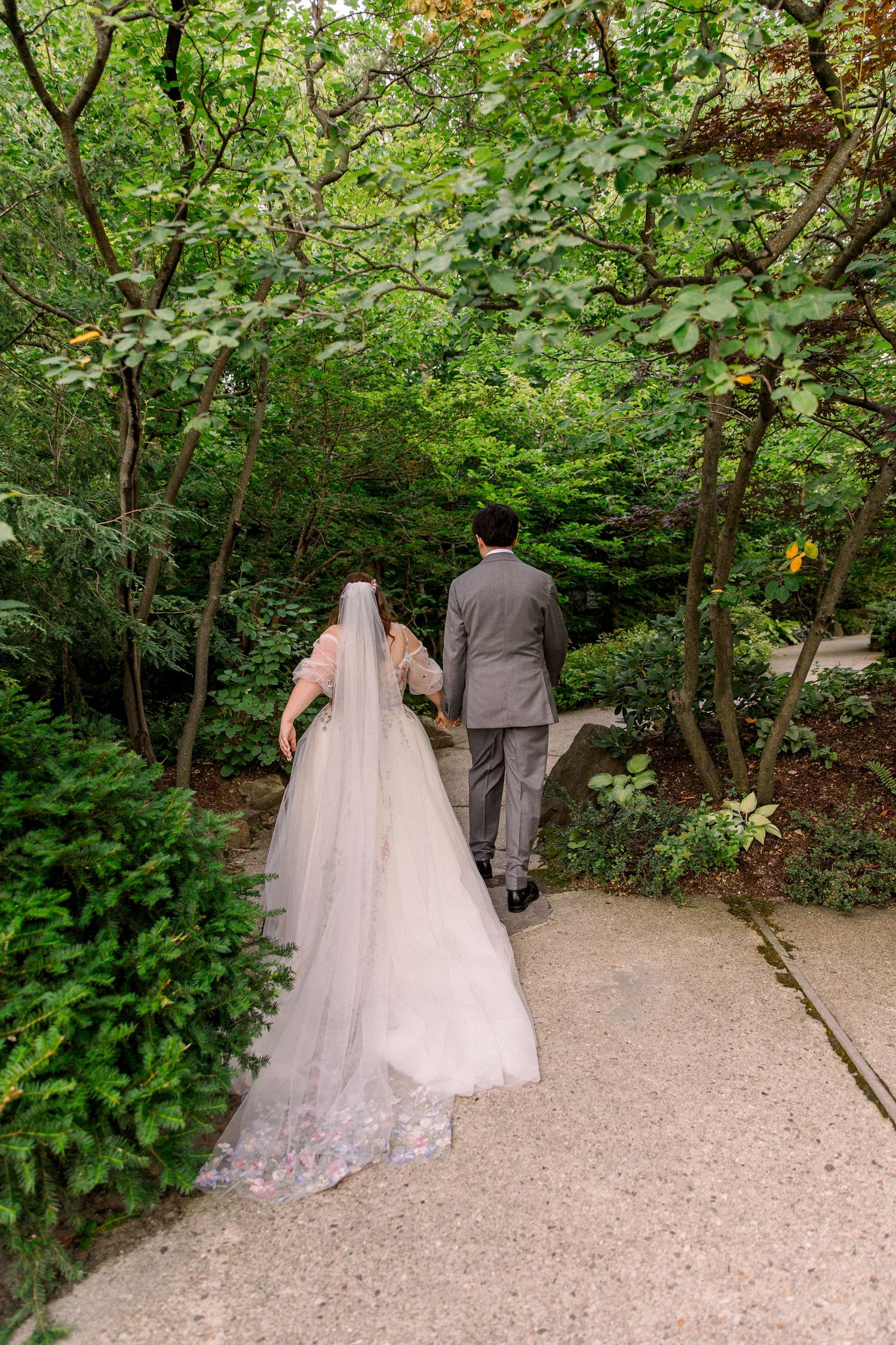 Bride and groom walking hand in hand through a lush green garden path, bride wearing a long white gown with veil.