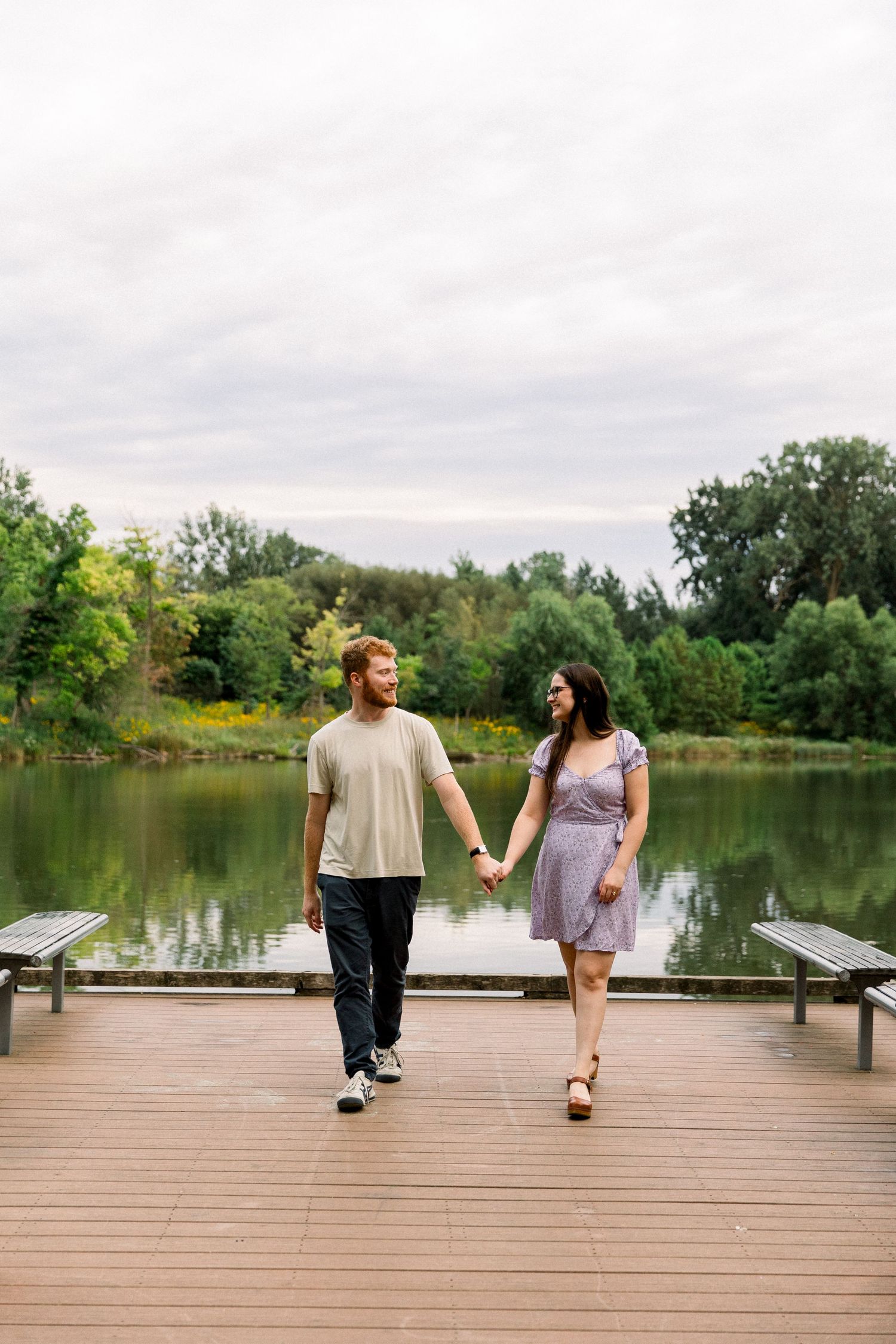 Couple holding hands walking on a wooden dock by a serene lake surrounded by lush green trees on a cloudy day.