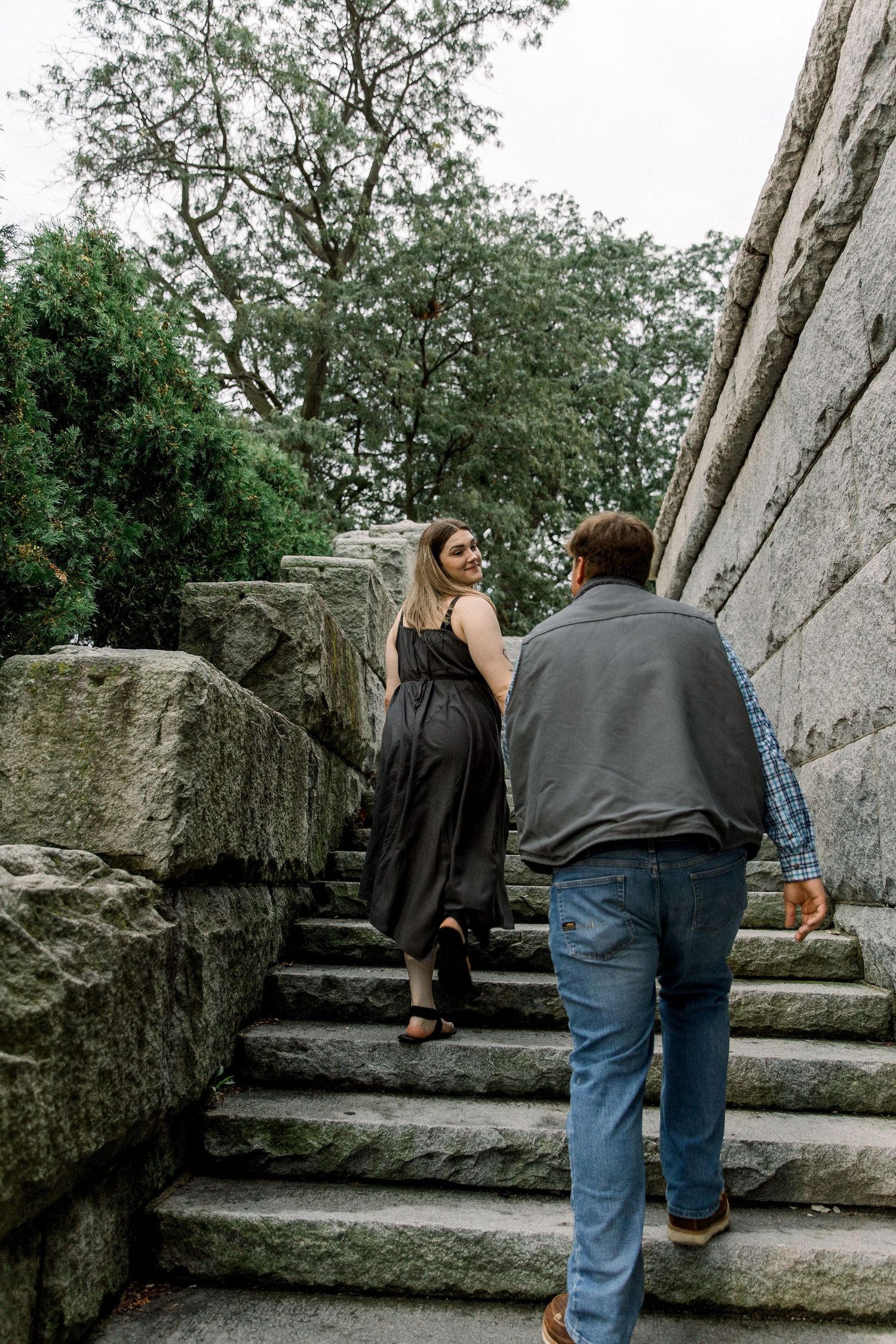Couple climbing stone stairs in a park, woman in black dress looks back smiling at man in gray vest and jeans.