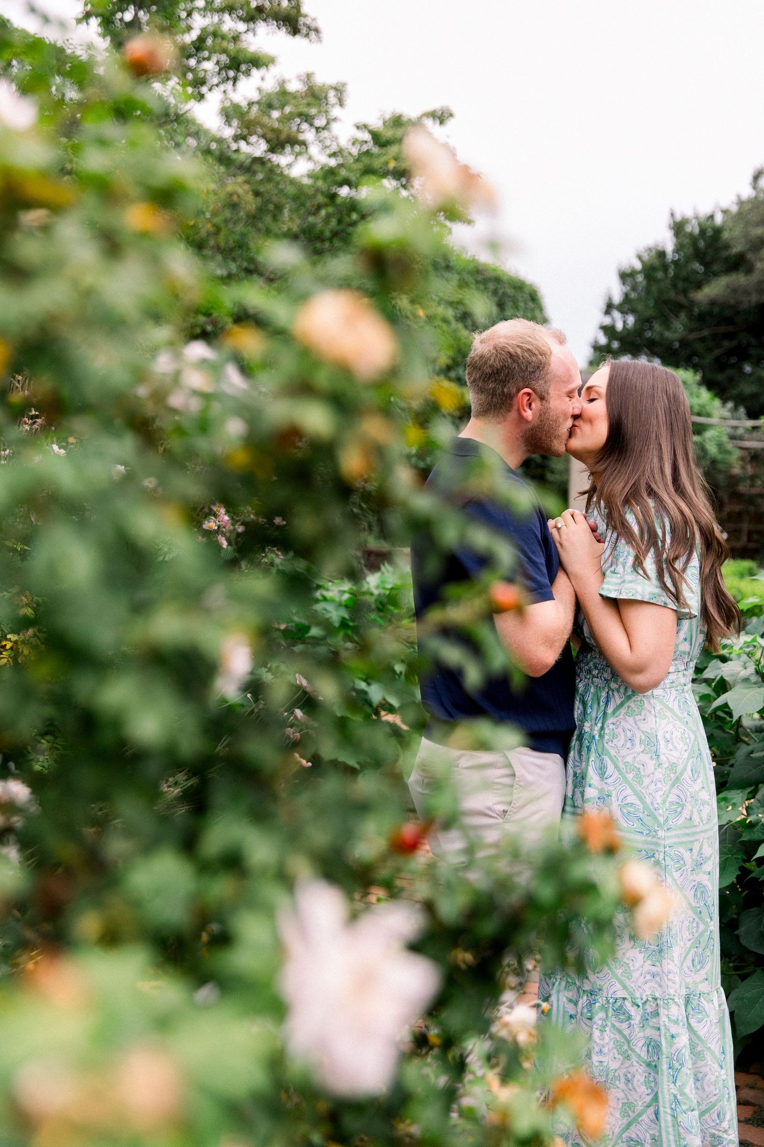 Couple sharing a romantic kiss surrounded by lush rose bushes in a garden.