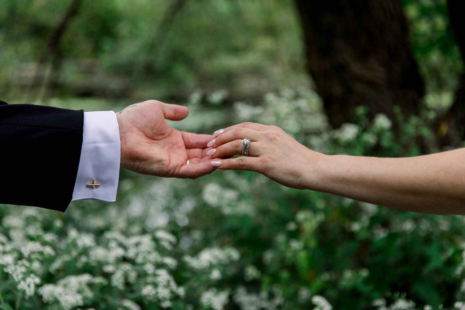 Bride and groom touching hands outdoors, showing wedding ring, with white wildflowers and green trees in background.