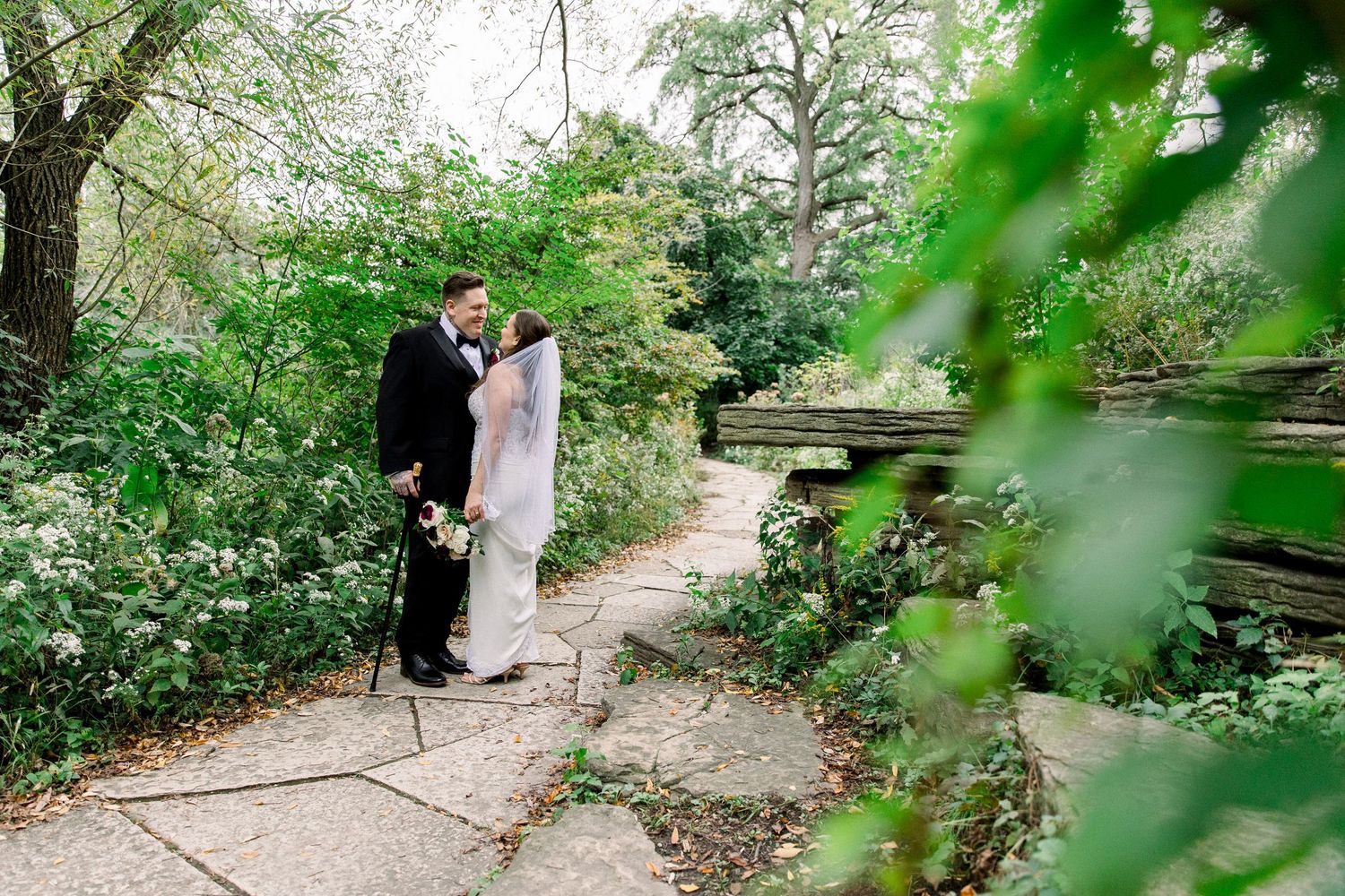 Bride and groom share a romantic moment in a lush garden setting, surrounded by green trees and stone pathways.