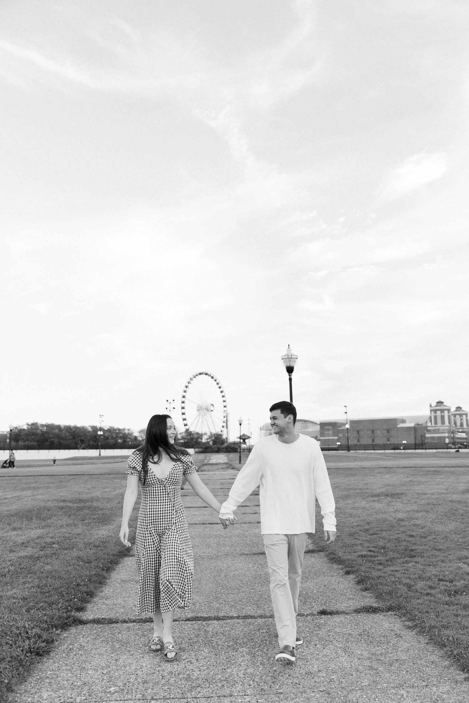 Black and white photo of couple holding hands and smiling at each other near a Ferris wheel outdoors.
