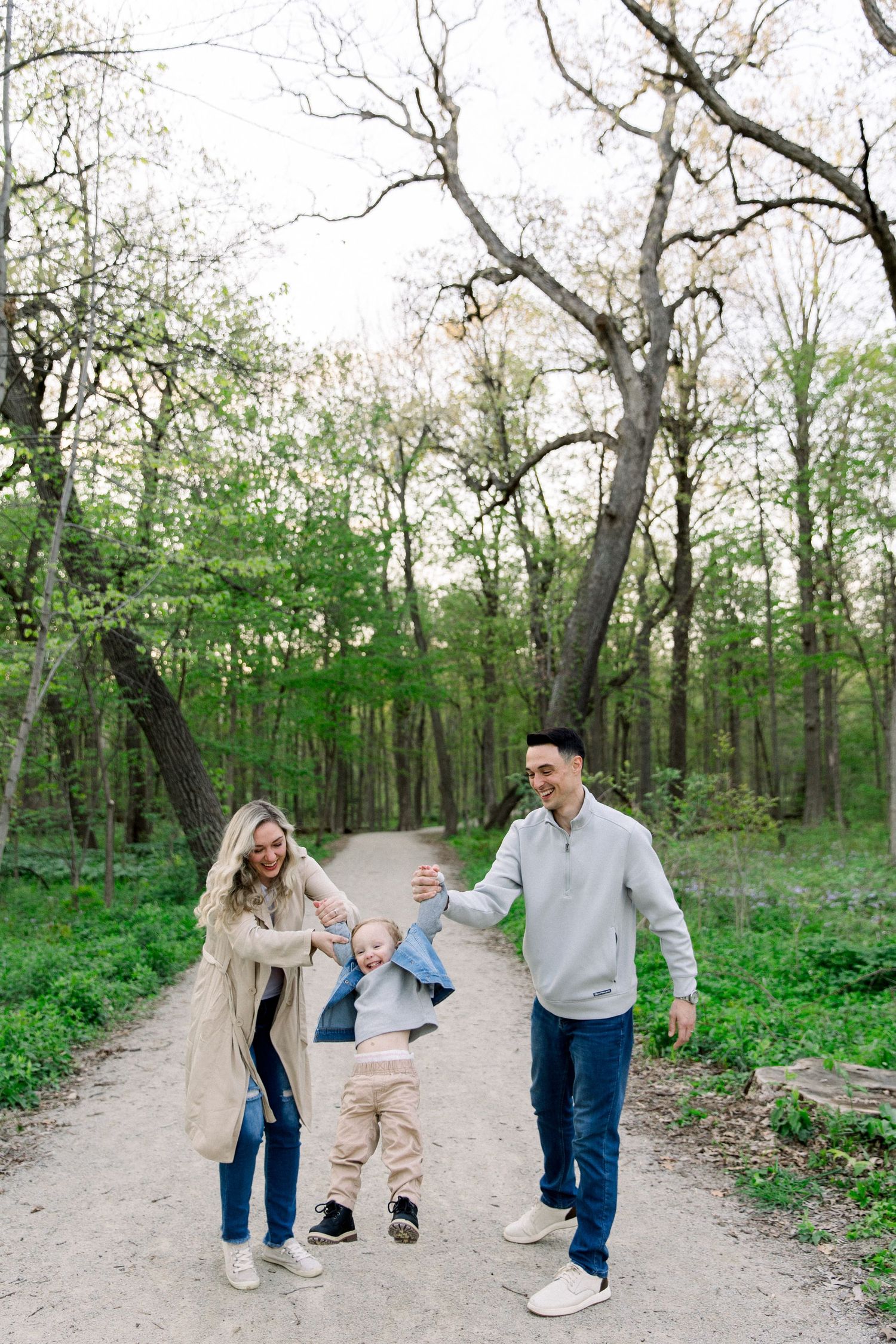 A happy family of three swings their toddler on a scenic wooded forest path in spring.