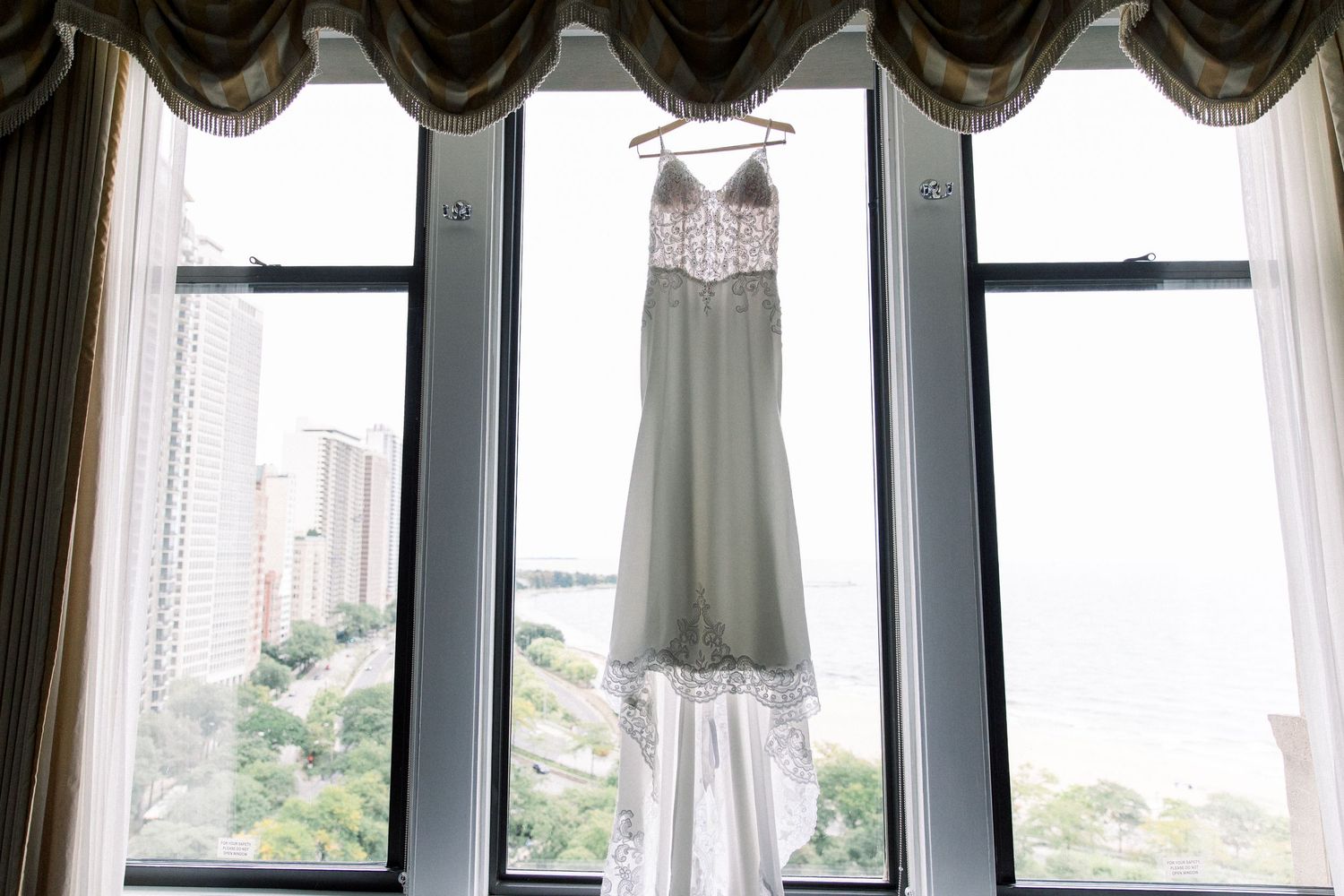 Elegant wedding dress hanging in a window with city skyline views on a bright day.