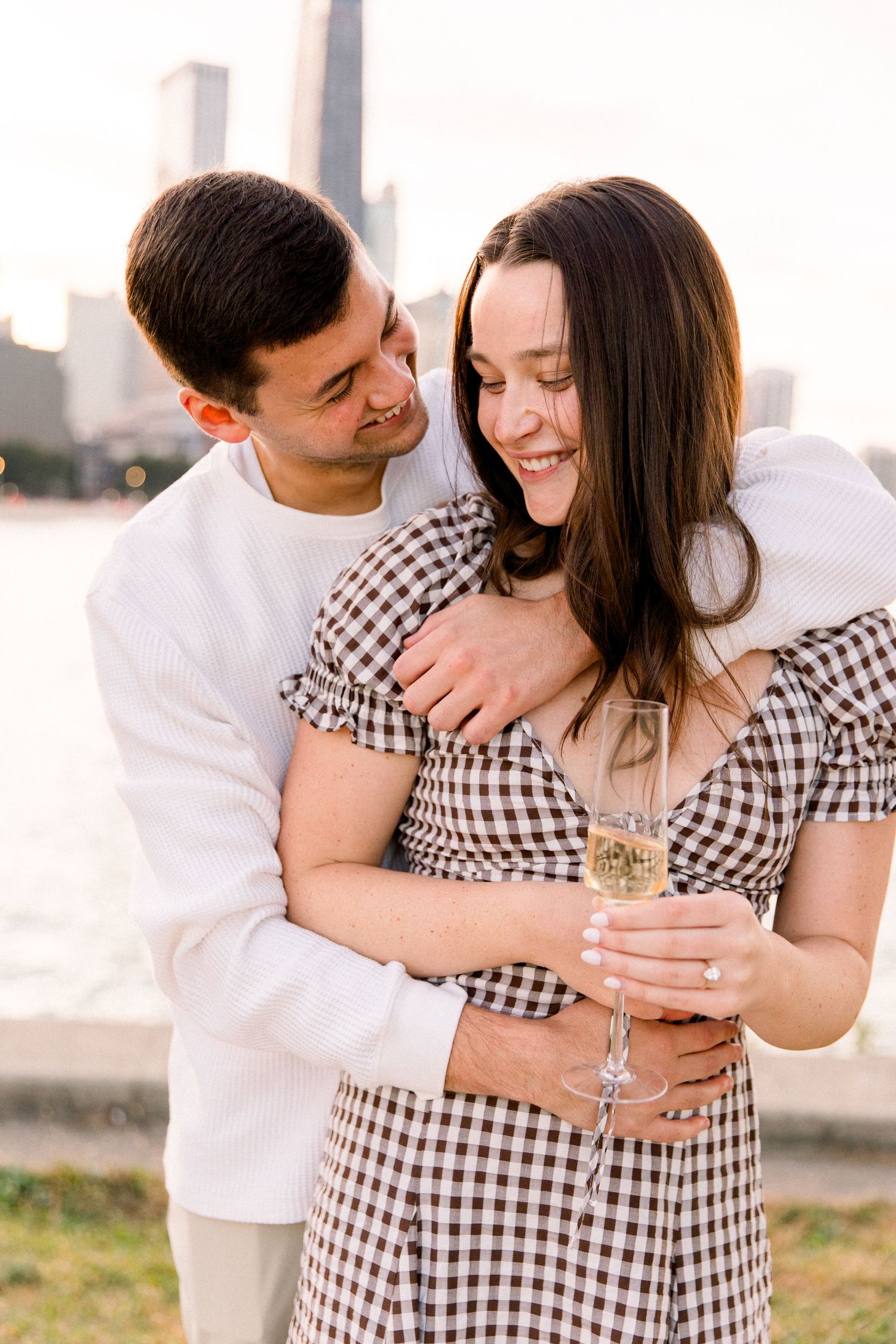 Happy couple embracing outdoors by waterfront, woman in plaid dress holding champagne glass, man in white shirt hugging her.