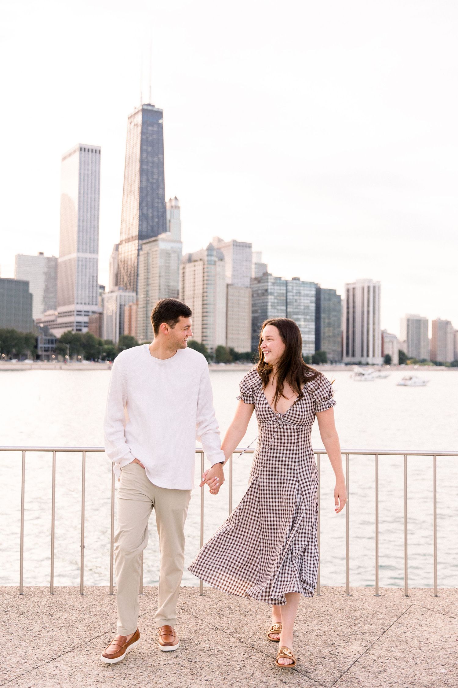 A couple holds hands and smiles while walking along a waterfront railing with the Chicago skyline behind them.