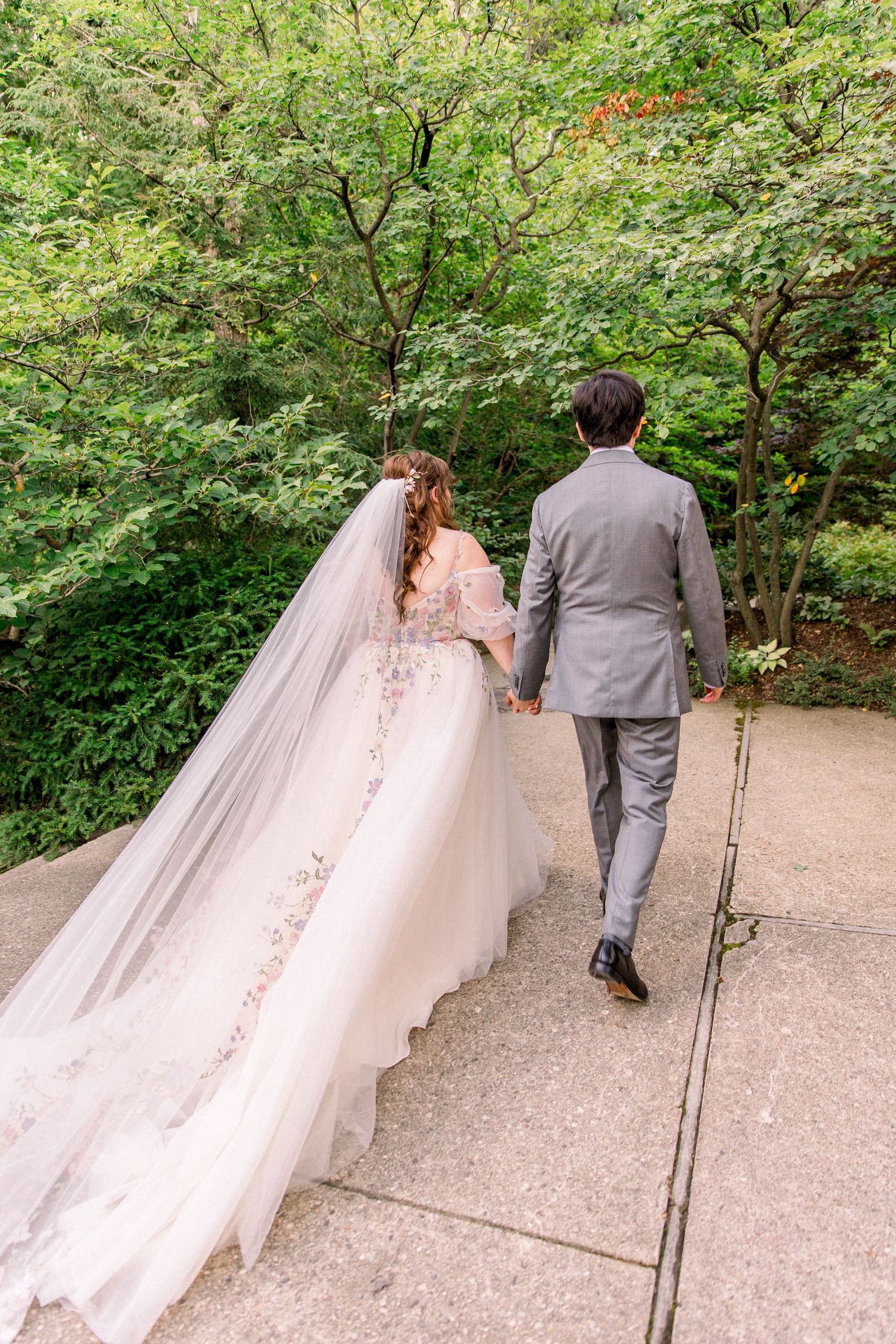 Bride and groom walking hand in hand up stone steps surrounded by lush green trees in a romantic outdoor wedding setting.