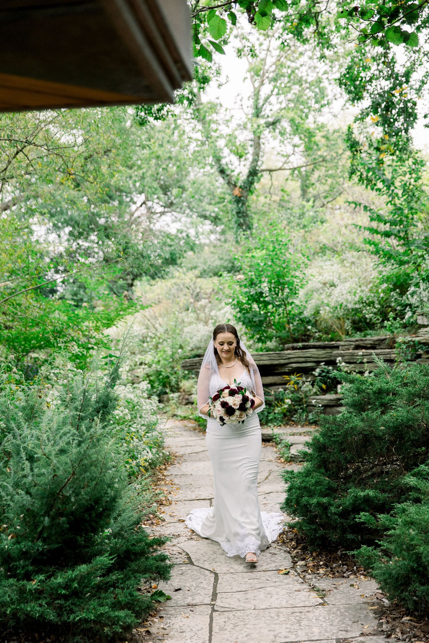 Bride in white gown and veil walks along stone garden path holding bouquet, surrounded by lush green trees.