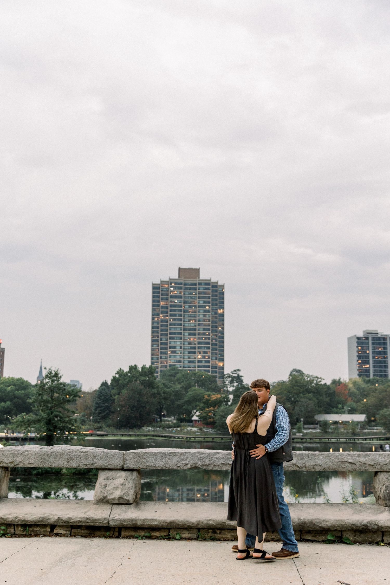 Couple embracing on a stone bridge overlooking a city skyline and lake on a cloudy day.