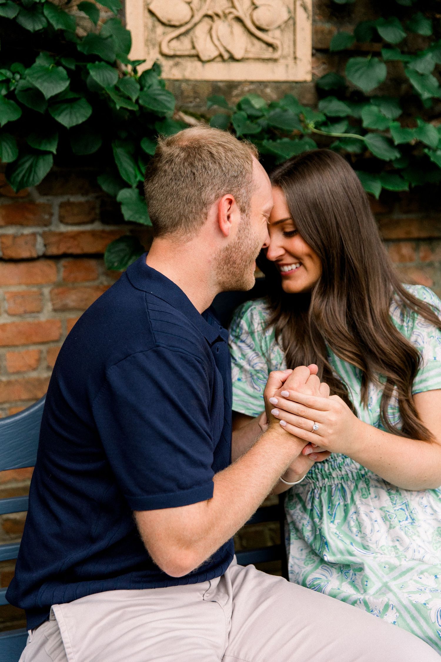 Couple sitting on bench holding hands and smiling at each other, woman wearing engagement ring, brick wall with ivy background.