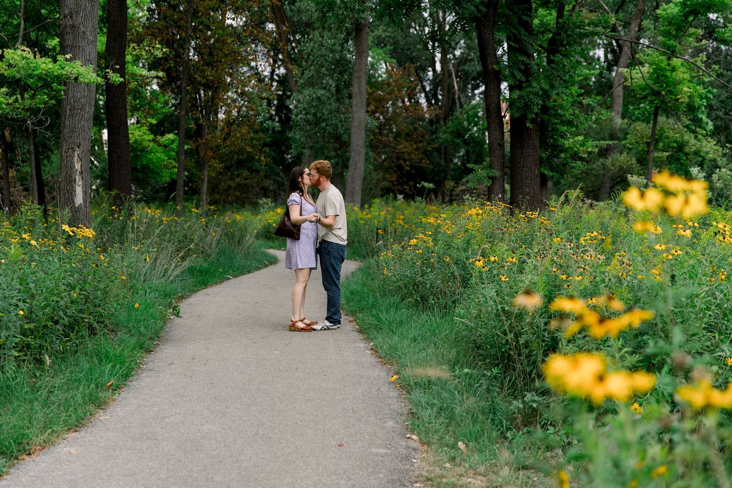 A couple shares a romantic moment on a scenic park path surrounded by lush green trees and vibrant yellow wildflowers.