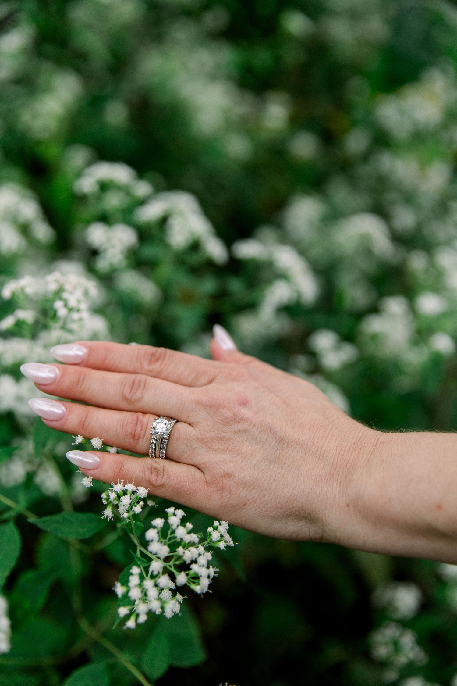 Close-up of a bride's hand displaying a diamond engagement ring and wedding band among white flowers.