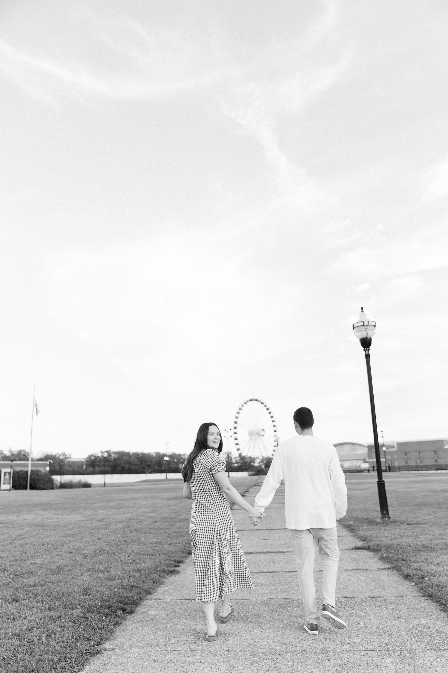 Black and white photo of couple holding hands walking toward Ferris wheel on Chicago's Navy Pier path.