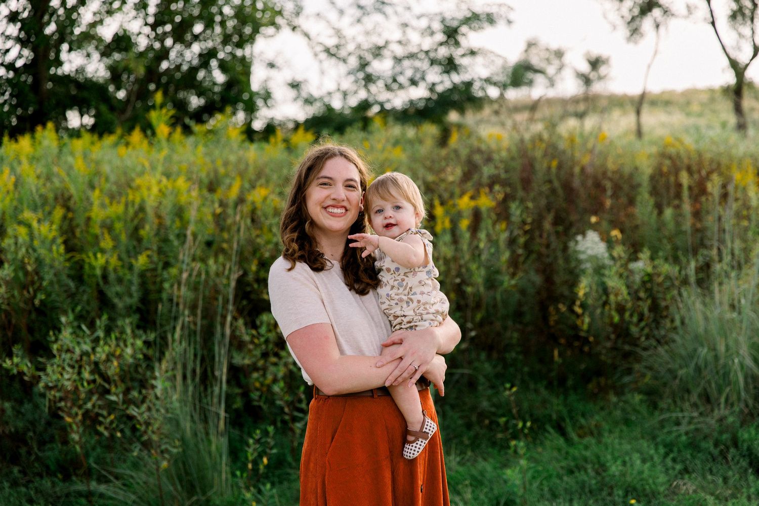A smiling woman holds a happy toddler in a sunlit field with wildflowers in the background.